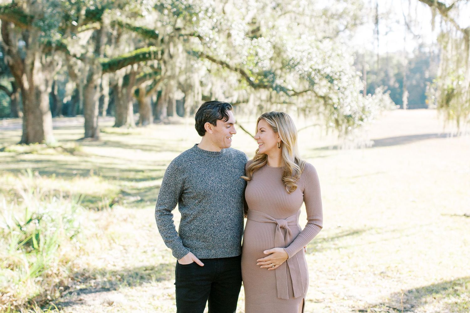 Romantic couple in casual sweaters stand together on a sunlit path beneath oak trees draped in Spanish moss.