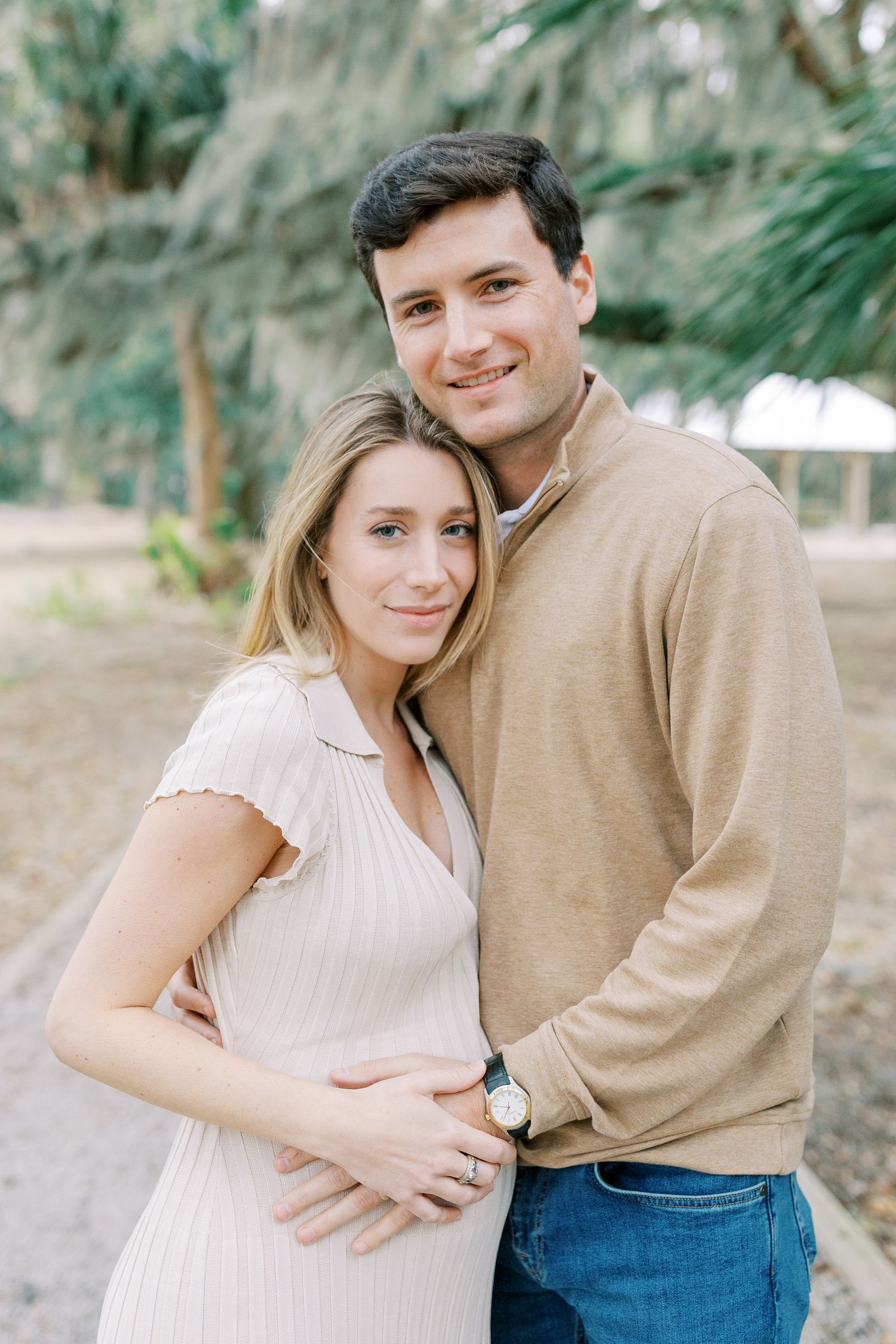 A couple in casual attire poses outdoors against a blurred natural backdrop on a sunny day.