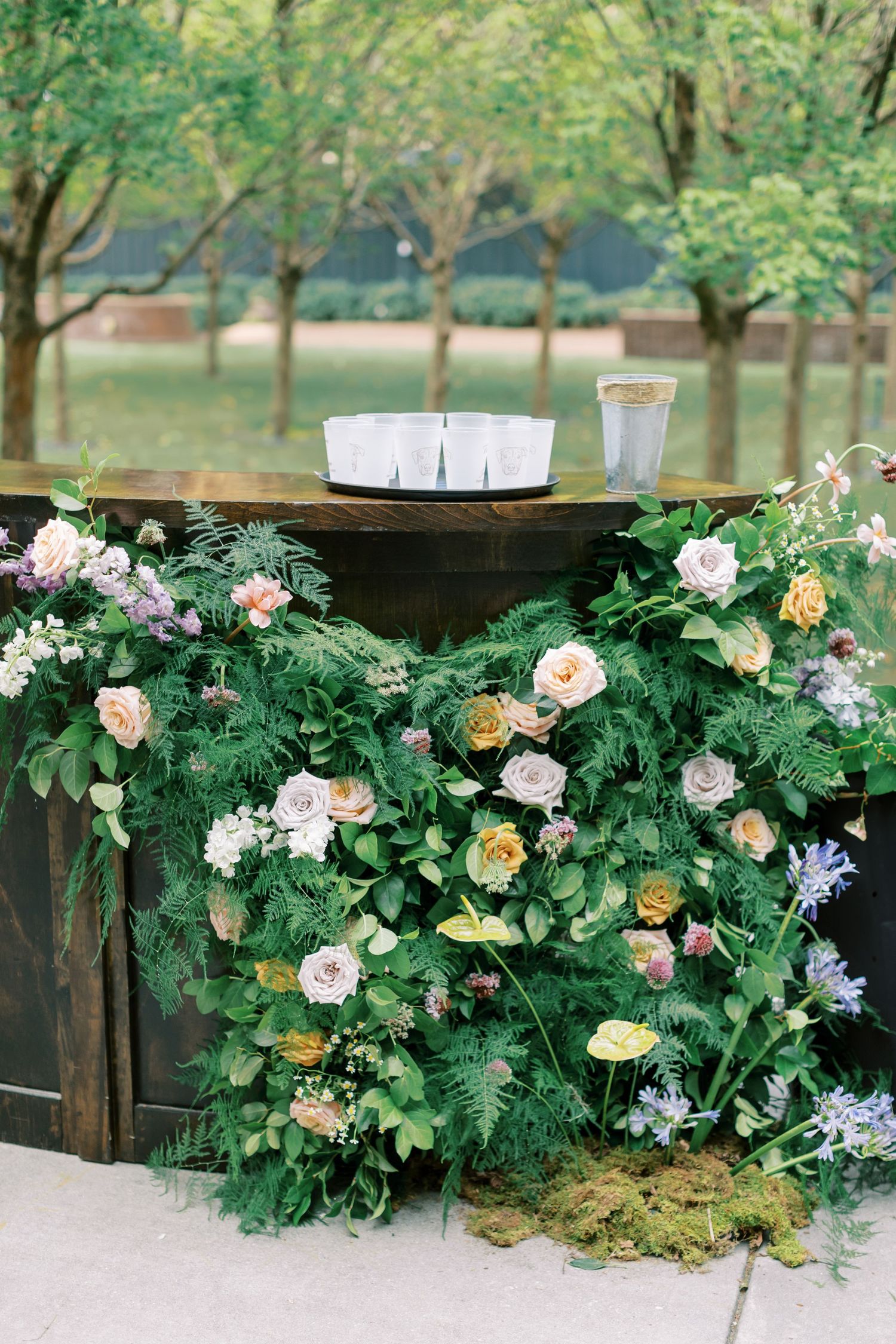 A rustic wooden bar decorated with lush greenery and pastel roses in an outdoor garden setting.