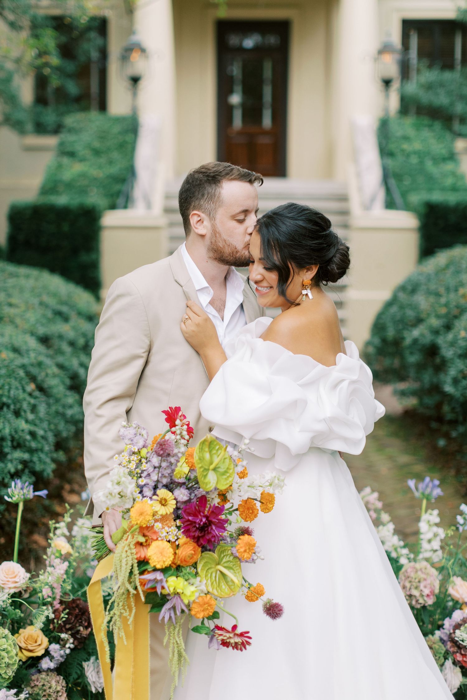 A romantic wedding couple shares a tender moment in a garden setting with a vibrant yellow and purple floral bouquet.
