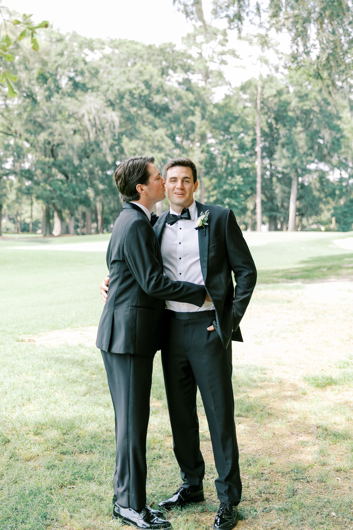A joyful wedding couple in formal attire share an intimate moment on lush grounds with hanging trees in background.