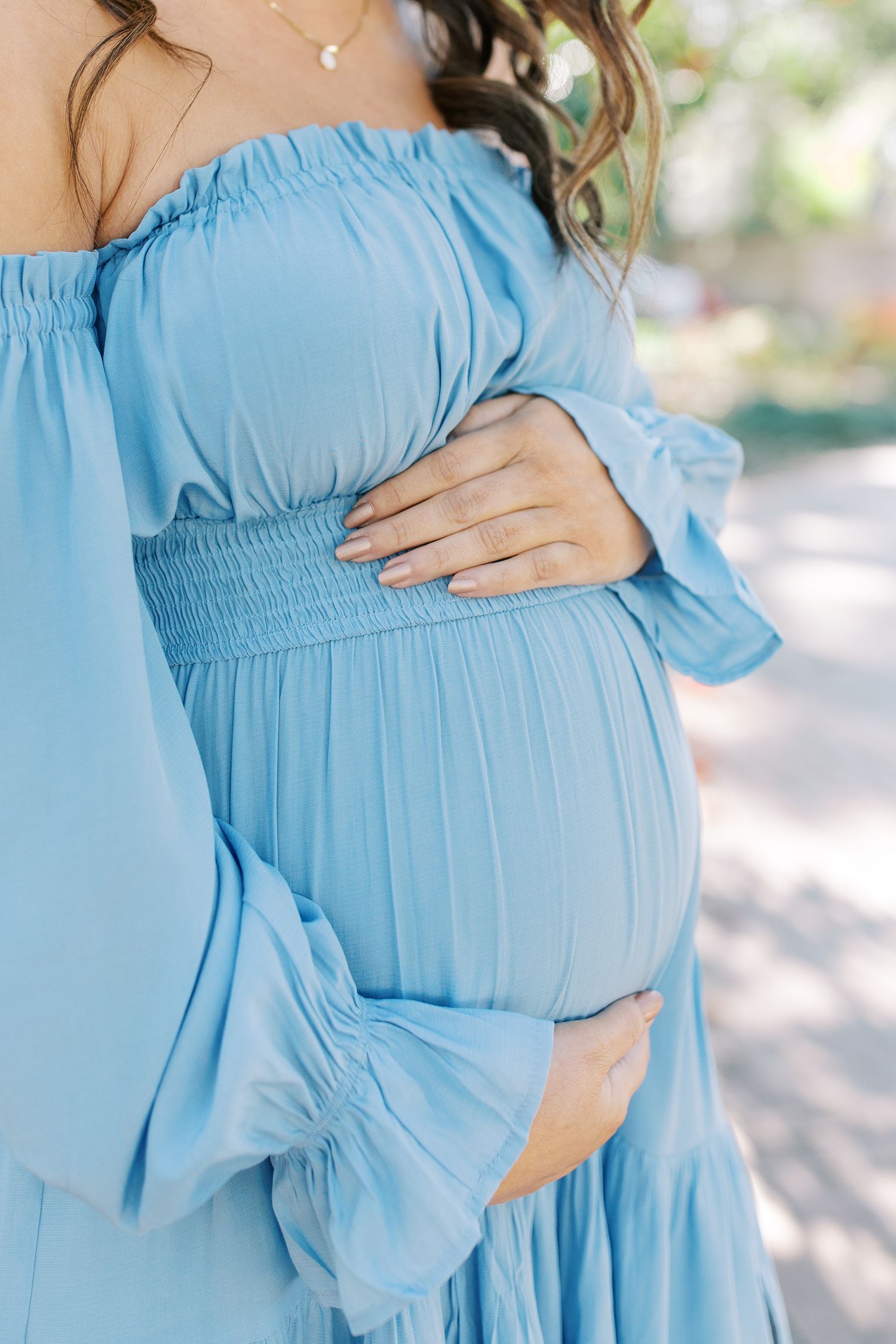 A close-up view of a pregnant belly in a flowing light blue off-shoulder maternity dress.