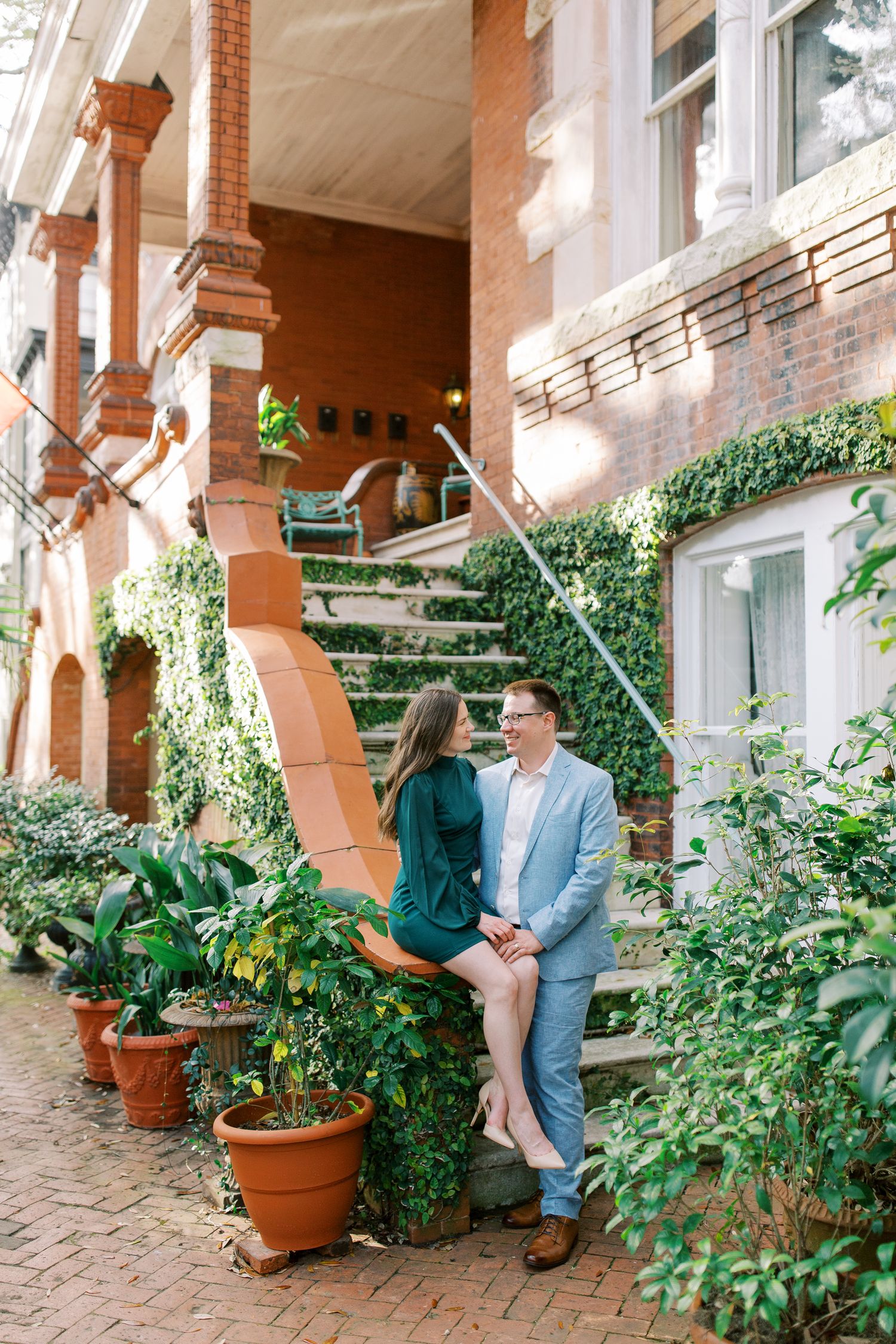 A couple sits together on brick steps surrounded by lush greenery and potted plants at a historic brownstone entrance.