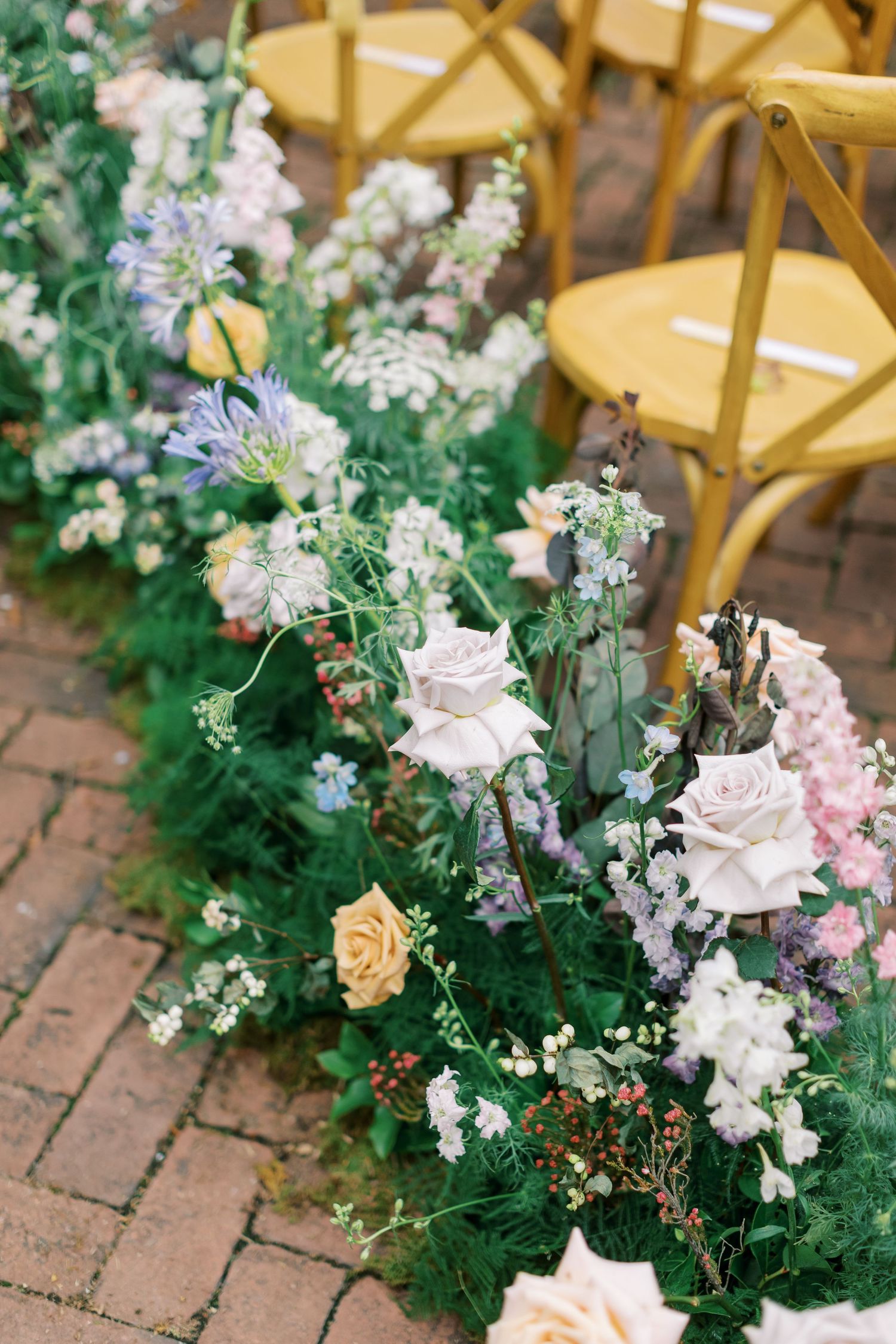 Delicate floral arrangements with pink roses and purple blooms line a brick pathway next to yellow wooden chairs.
