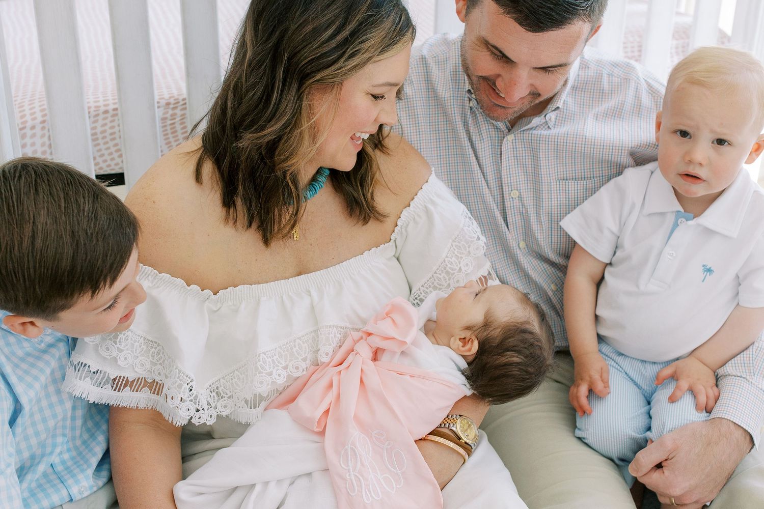 Family shares tender moment with newborn sibling in bright white nursery setting.