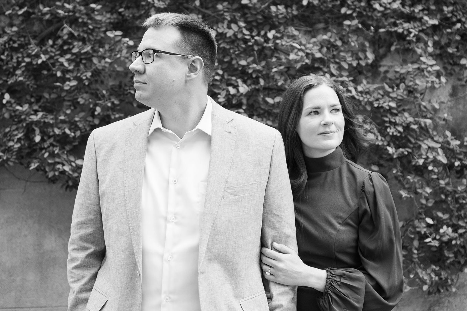 A black and white portrait of a couple outdoors looking in opposite directions, with greenery in the background.
