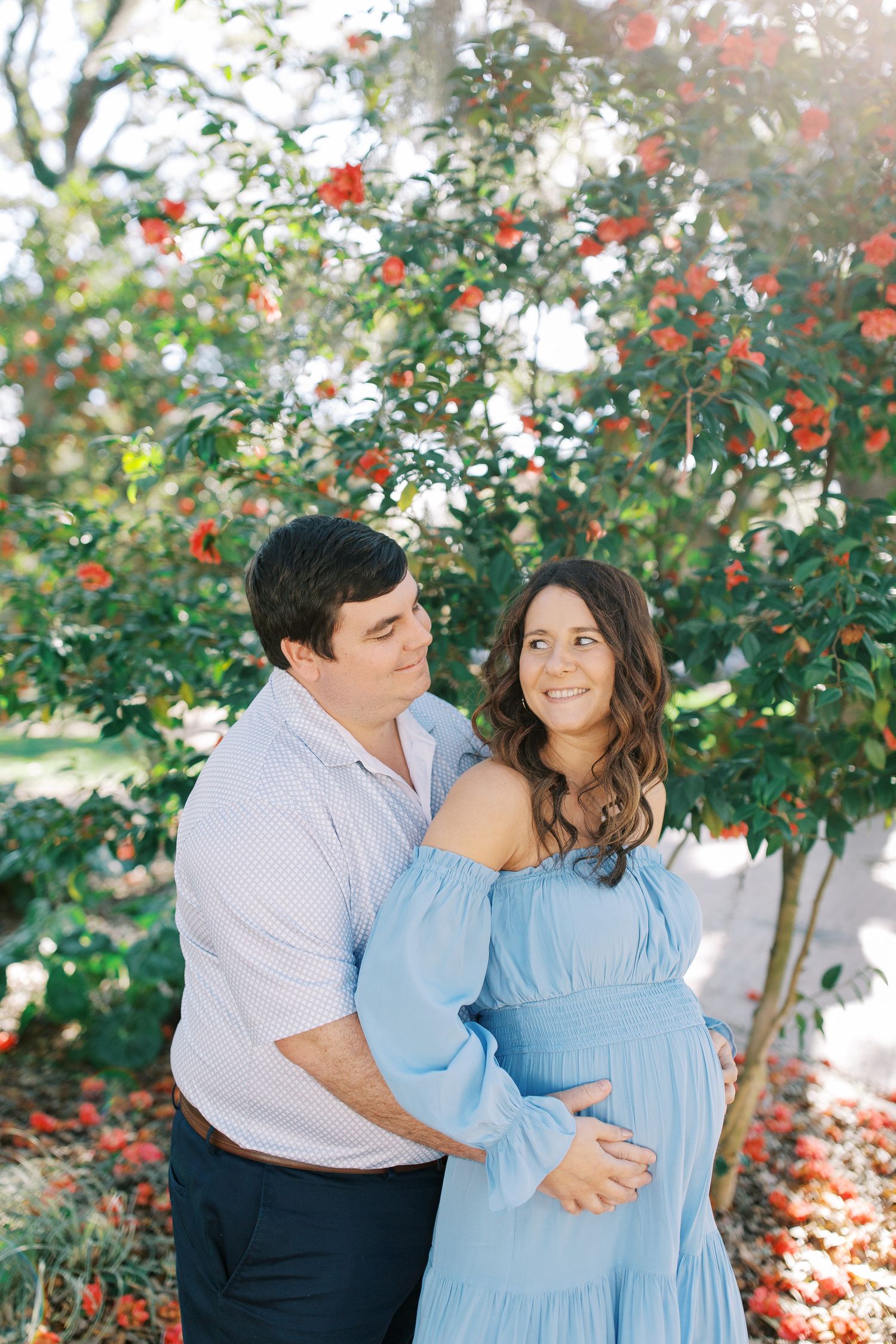 A couple poses together near blooming red flowers, with soft sunlight filtering through the trees.