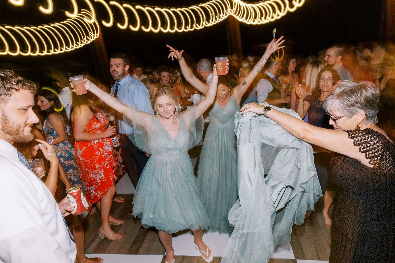 Wedding party celebrating on dance floor under twinkle lights with guests in pastel dresses.