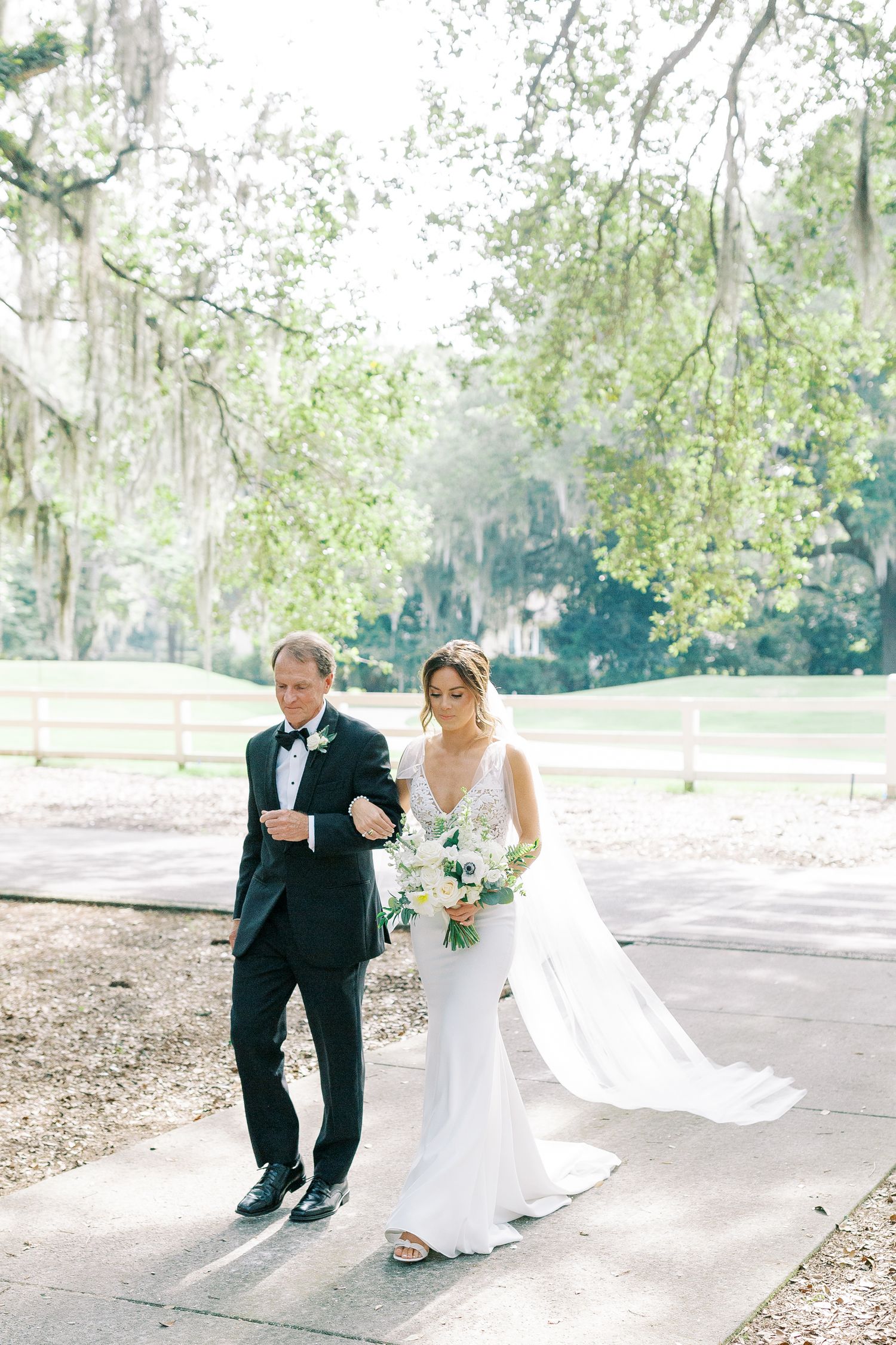 Bride and her father walk arm in arm on a garden path under Spanish moss-draped trees during their wedding celebration.