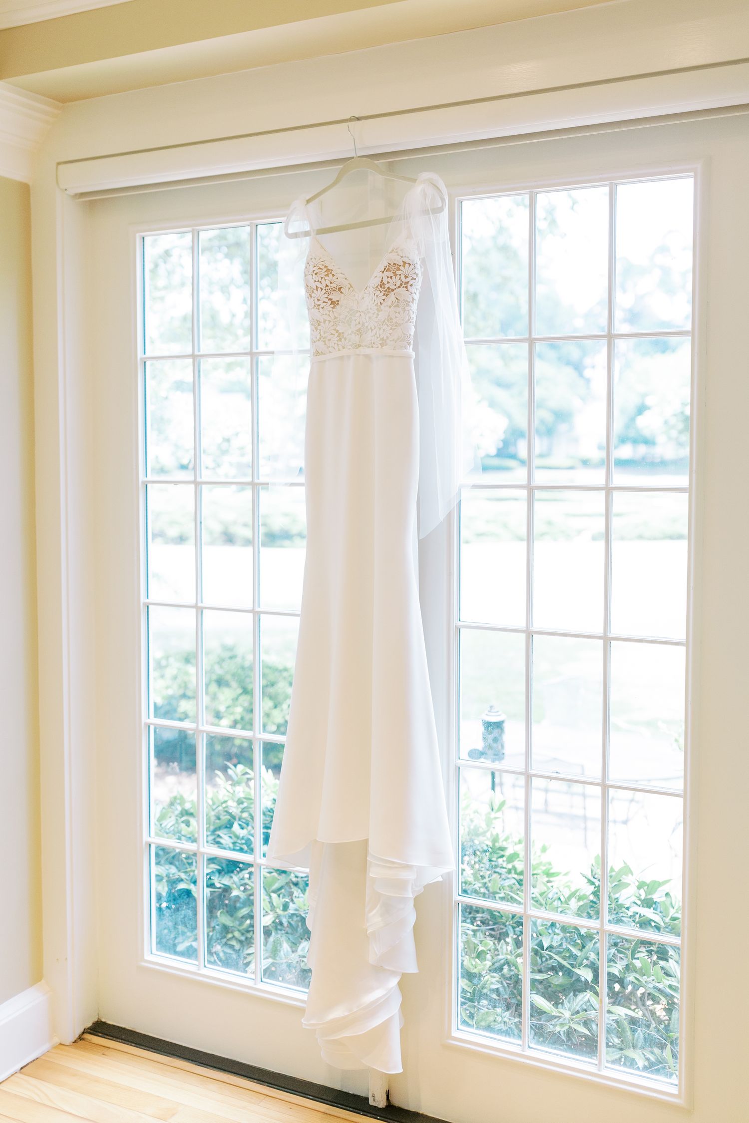 Elegant white wedding dress with beaded bodice hangs gracefully in front of bright floor-to-ceiling windows.