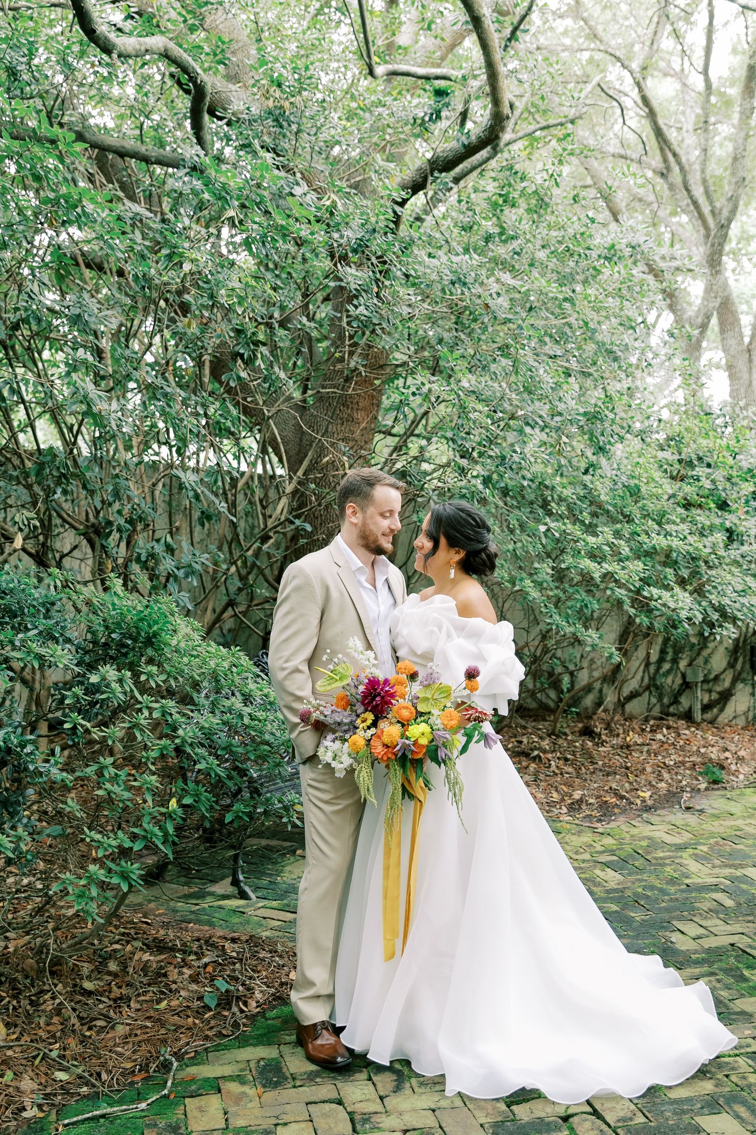 Newlyweds pose with colorful bouquet along garden path surrounded by lush greenery.