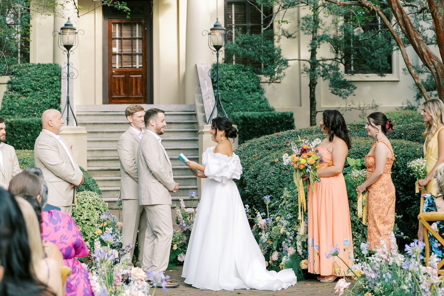 Outdoor wedding ceremony in progress surrounded by manicured gardens and peach-colored bridesmaid dresses.