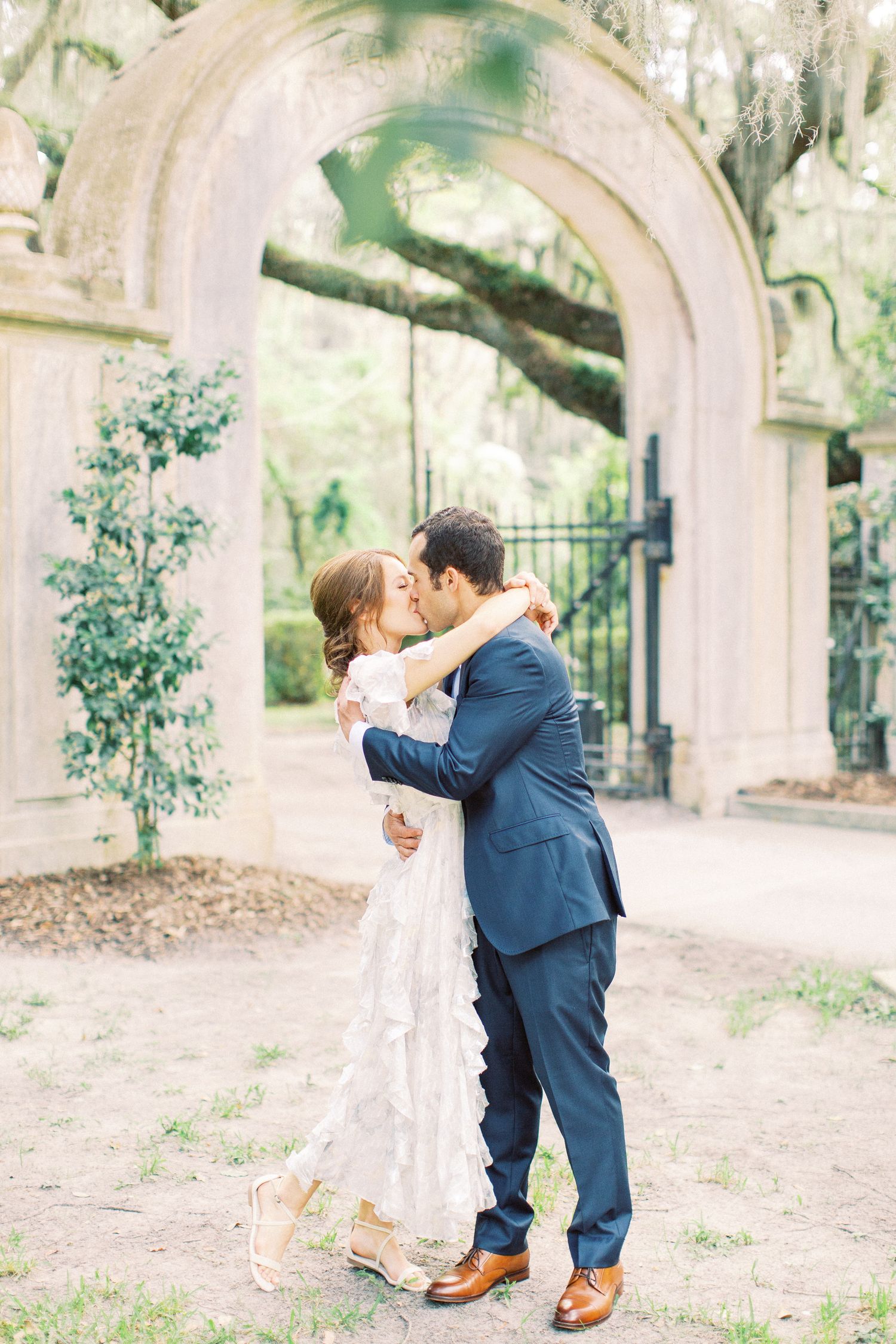 A couple in wedding attire share a romantic kiss under a stone archway in a garden setting at Wormsloe in Savannah, GA.