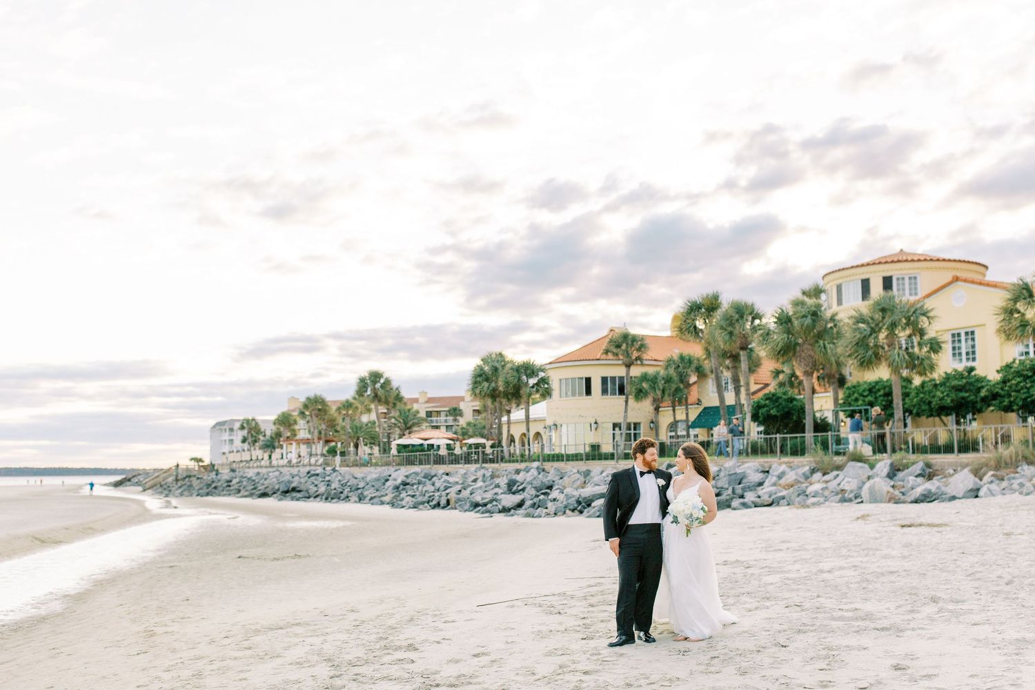 A couple shares a romantic moment on a sandy beach with coastal homes and palm trees in the background in St. Simons, GA.
