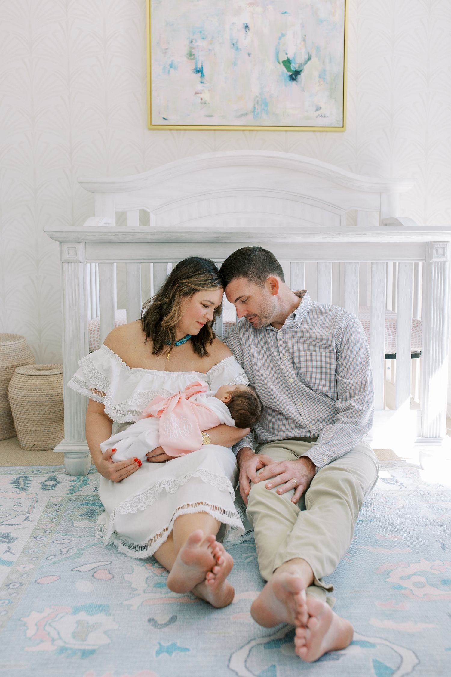 A couple sits barefoot on the floor next to a white crib, cradling their newborn baby in a nursery with blue decor.
