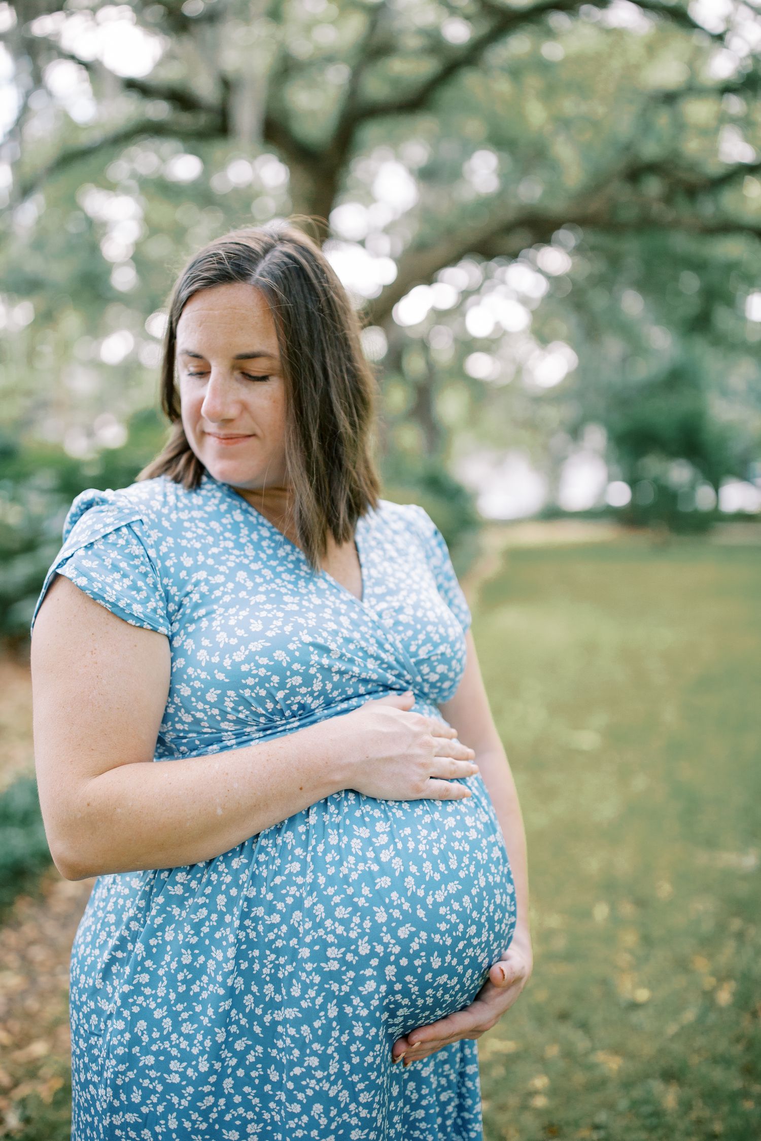 A person in a blue floral dress embraces their pregnant belly while standing outdoors near trees in forsyth park.