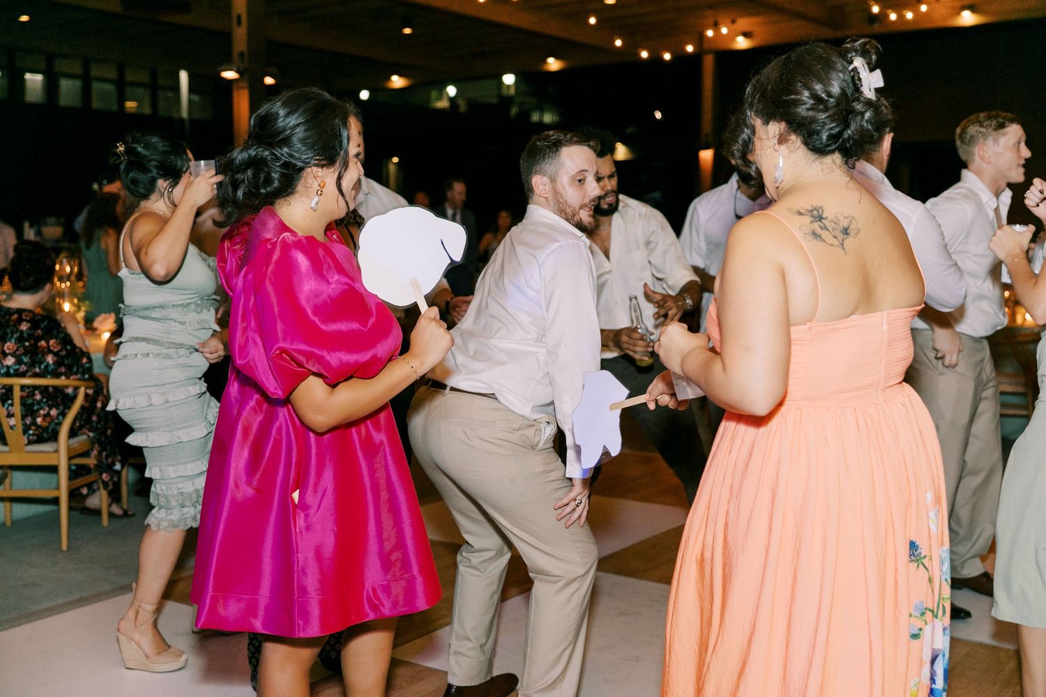 Wedding reception guests dancing and celebrating in a festive indoor venue with string lights.