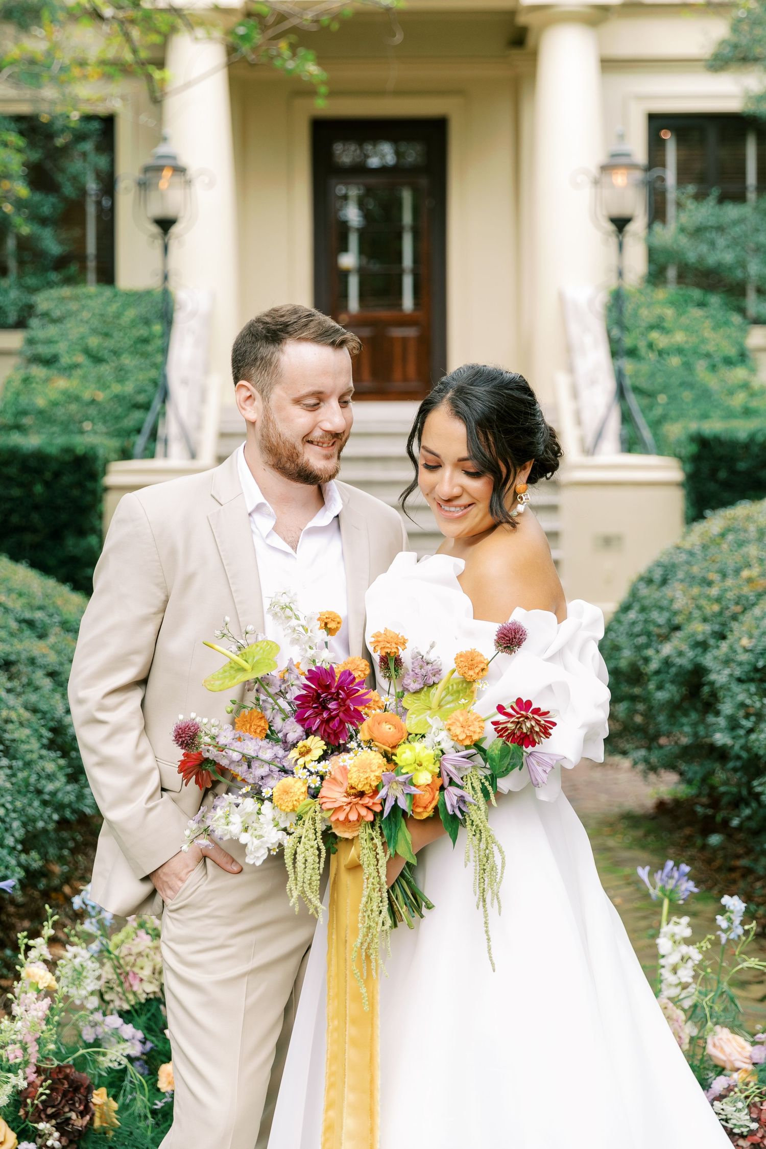 A couple shares romantic moments in front of a classic building while holding vibrant floral bouquets.