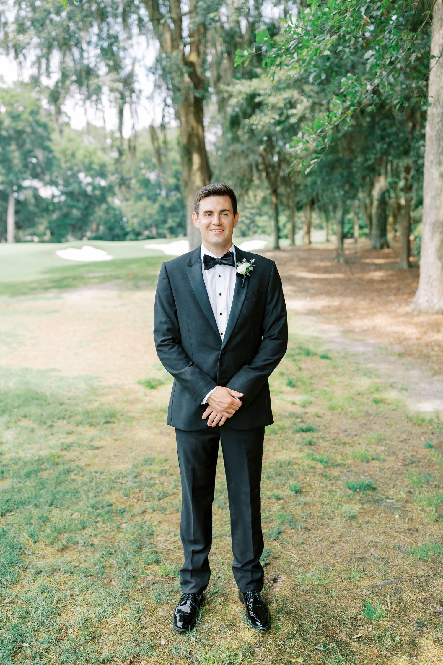 A groom in a formal black tuxedo and bow tie stands on grass beneath tall trees in an outdoor setting.