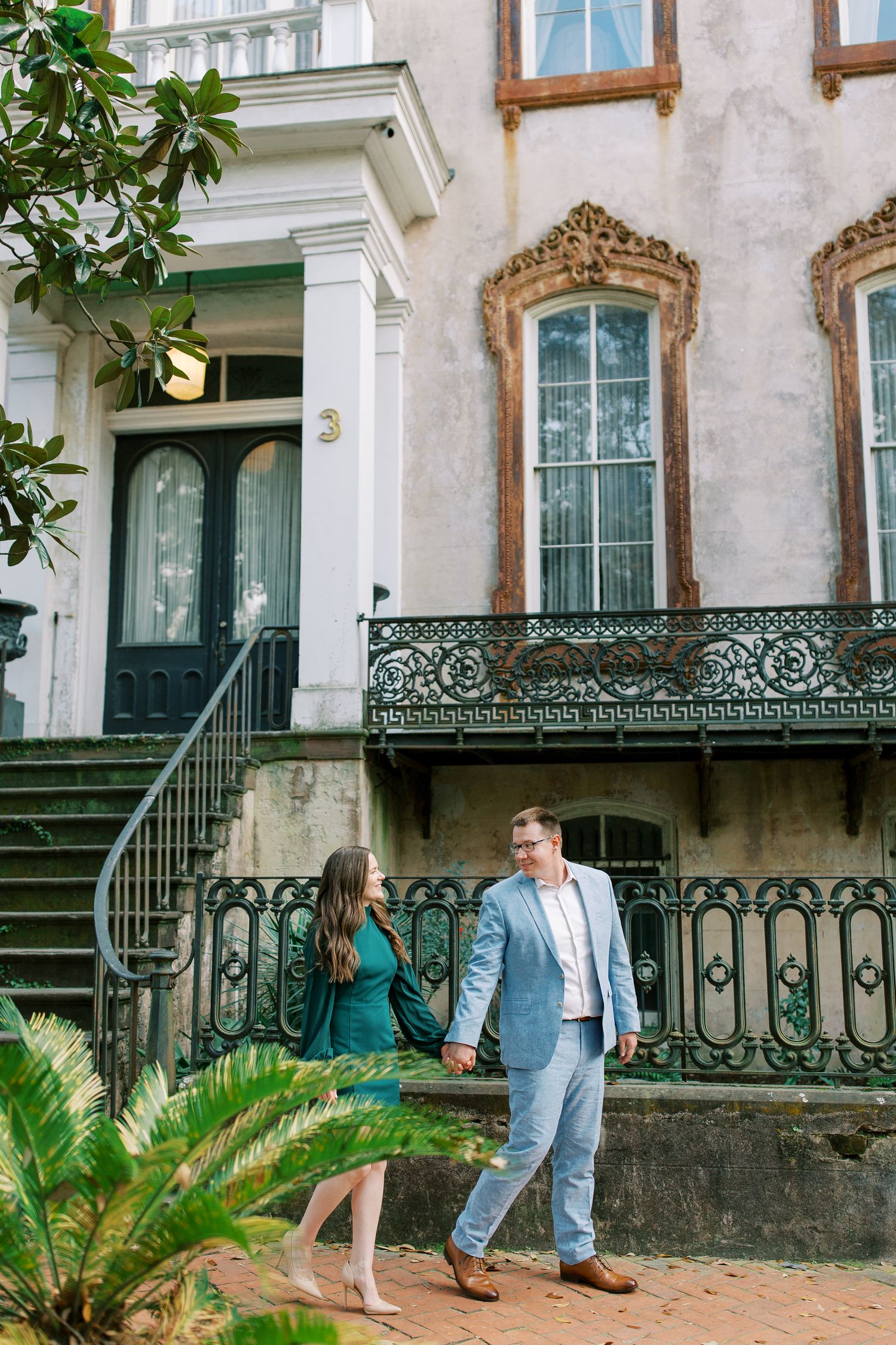 A couple stands on historic steps near an ornate iron balcony and architectural details.