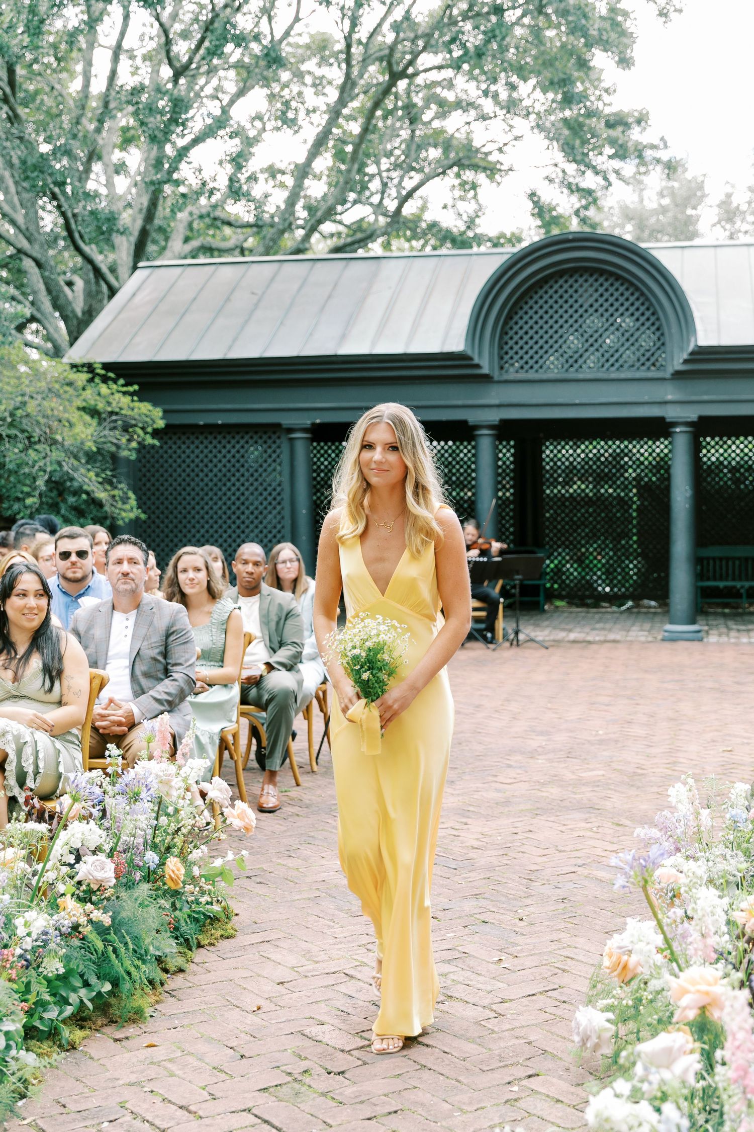 Wedding guests in bright summer attire walk along a landscaped brick path with green lattice pergola.