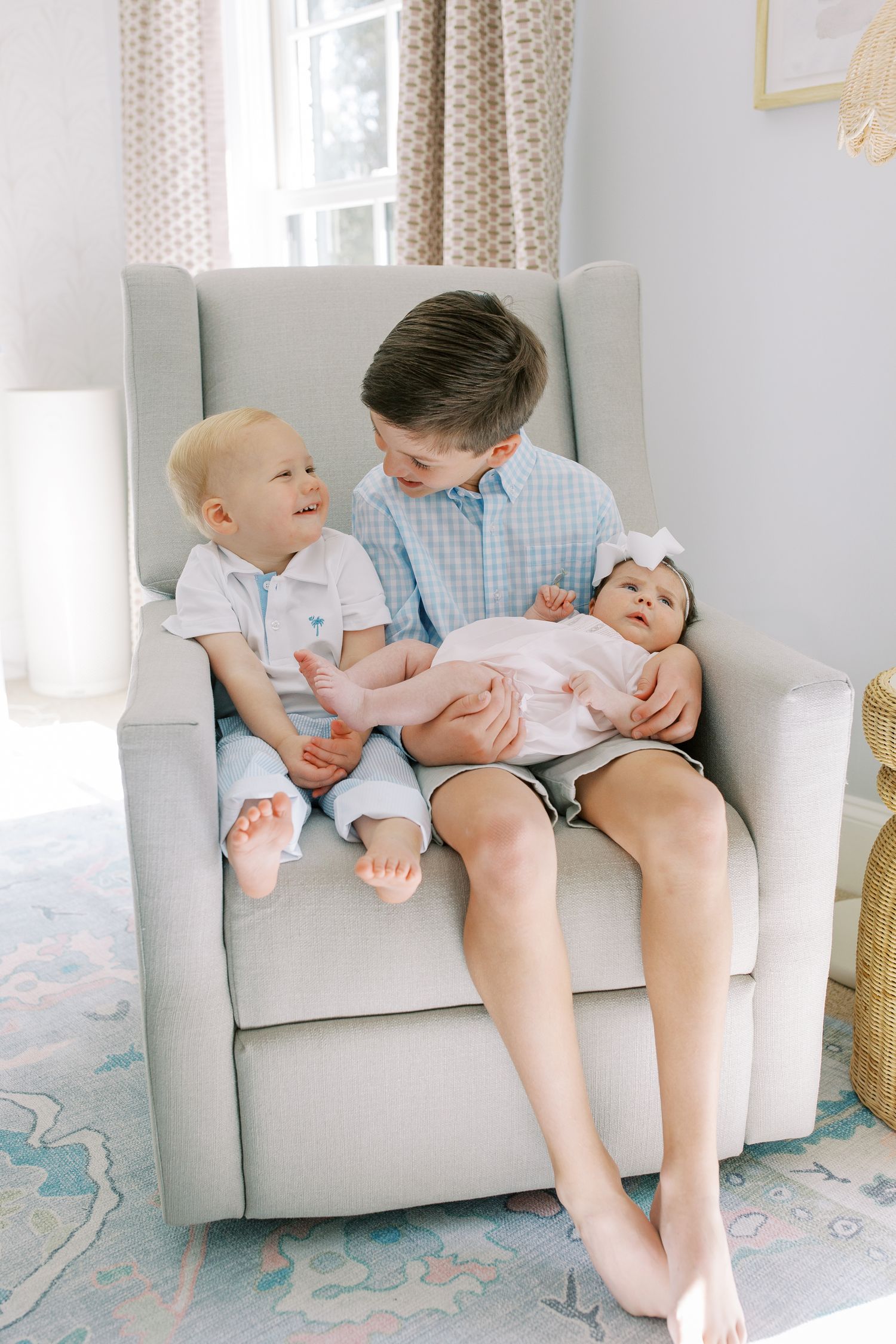 Three siblings share a sweet moment together in a gray armchair by a window.