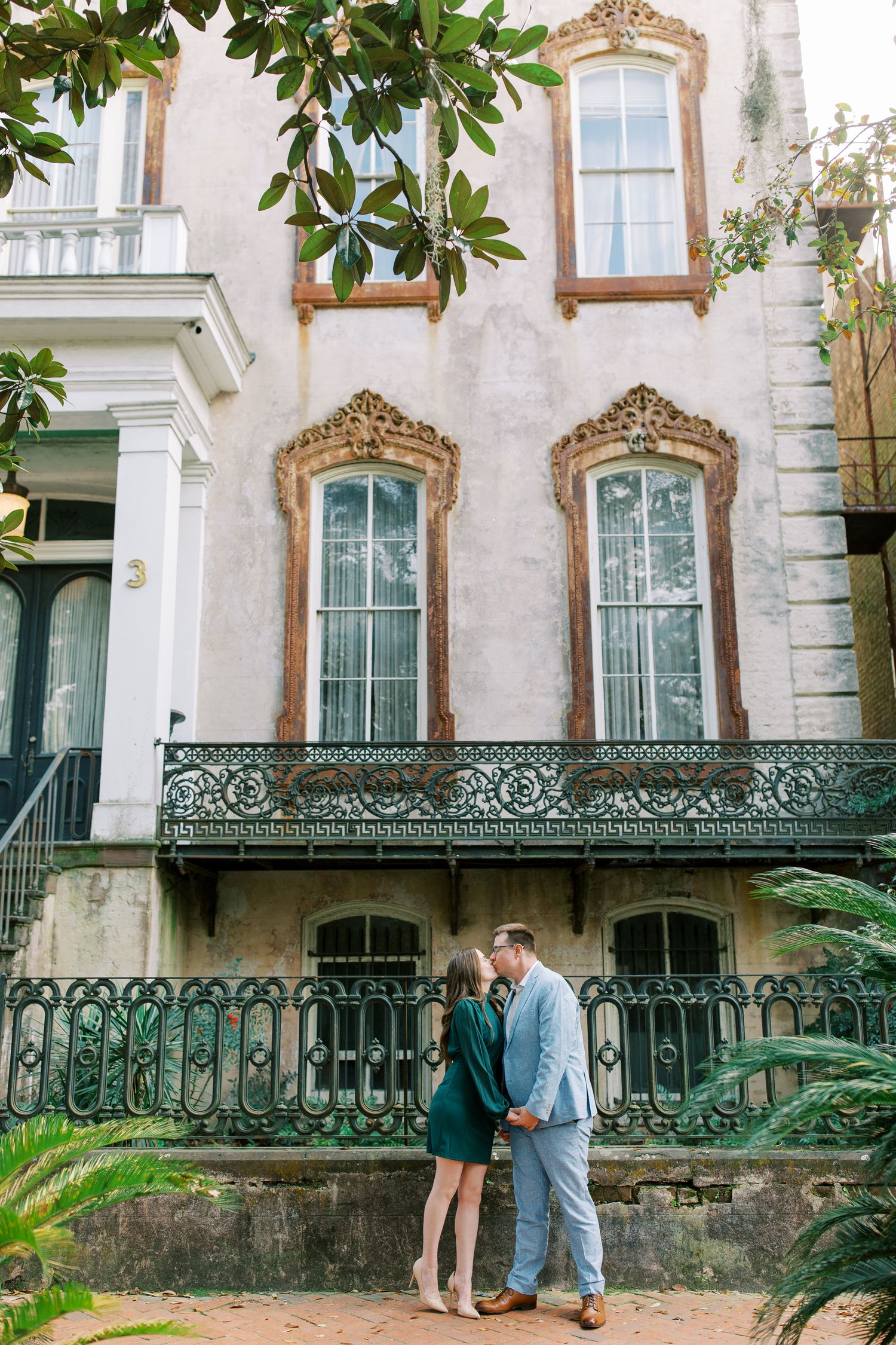 Ornate historic building in Savannah with a decorative wrought iron balcony and Victorian windows framed by lush greenery.