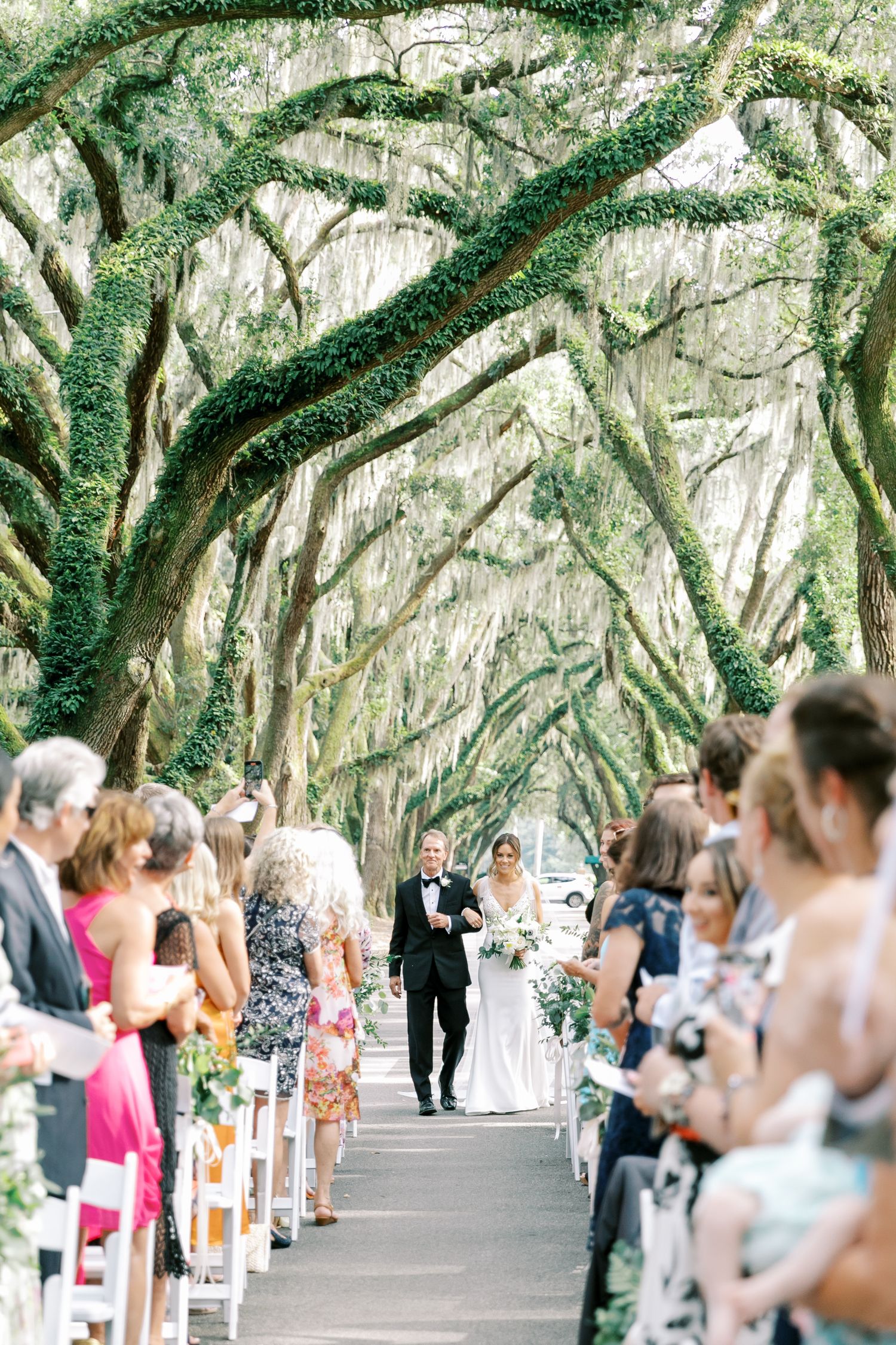 Couple walks down a tree-lined pathway during an outdoor wedding ceremony with guests seated on white chairs.
