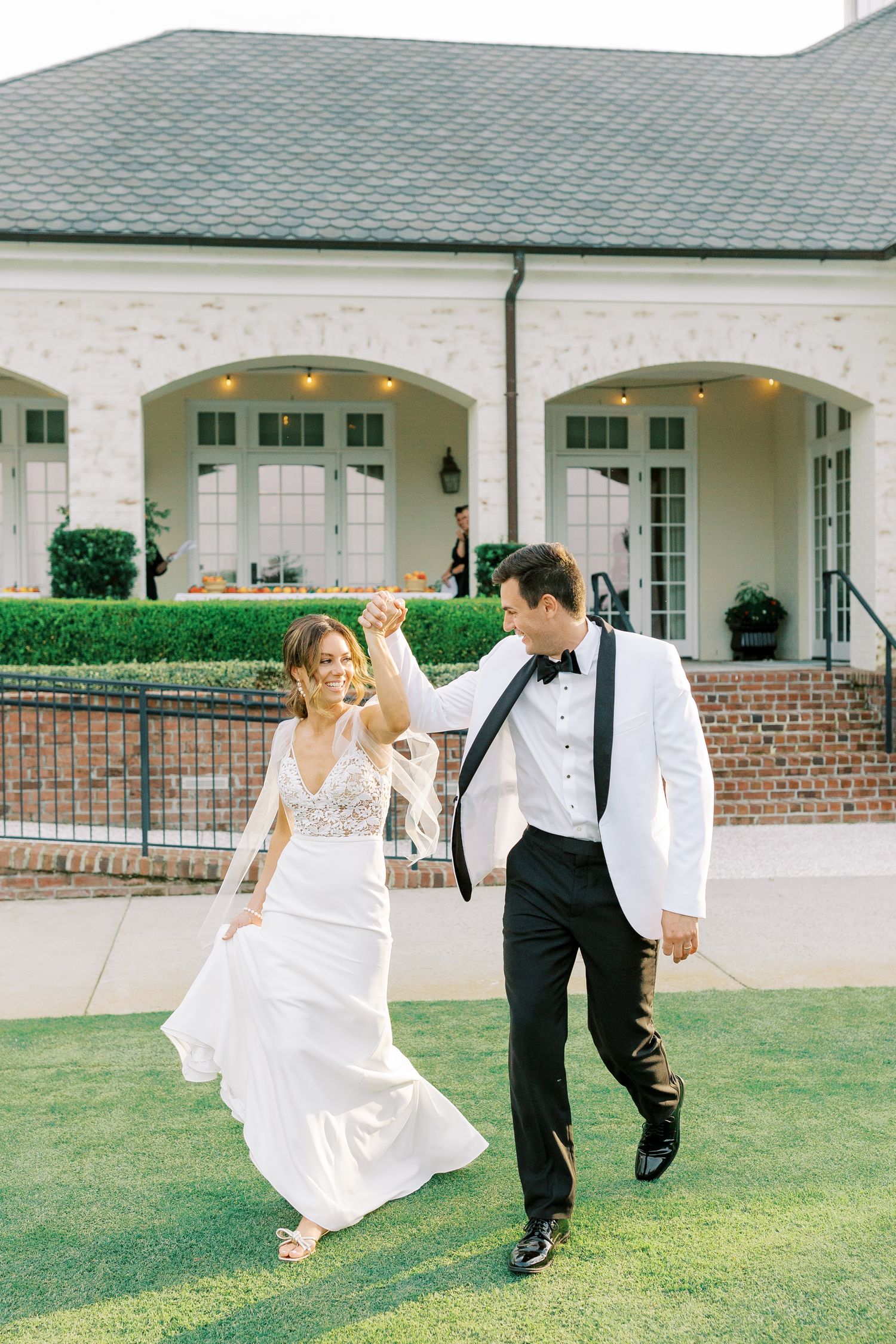 Newlyweds dancing joyfully on a manicured lawn in front of an elegant white house with green roof and arched doorways.