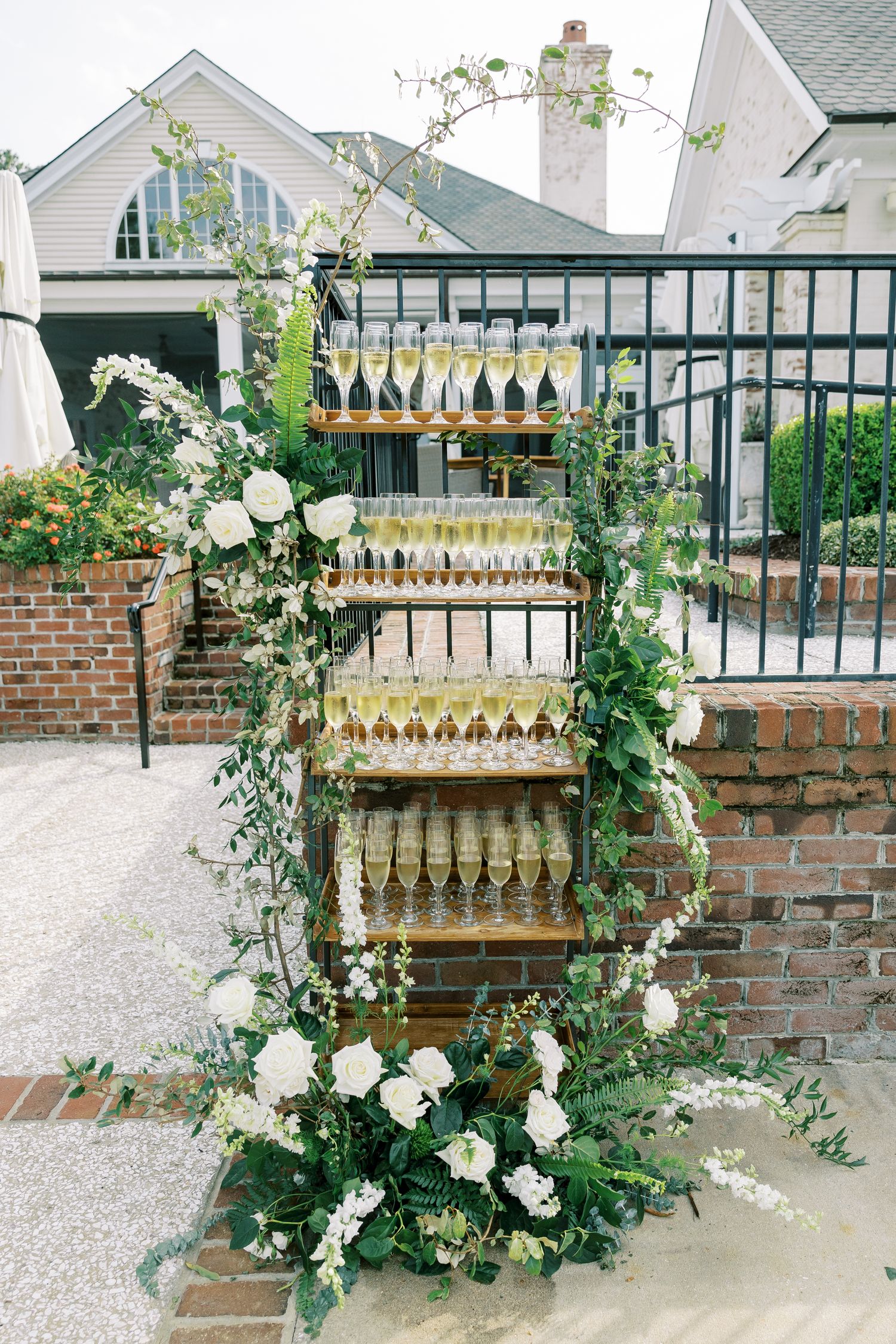 Elegant champagne wall display adorned with white roses and greenery vines against a brick and white house exterior.