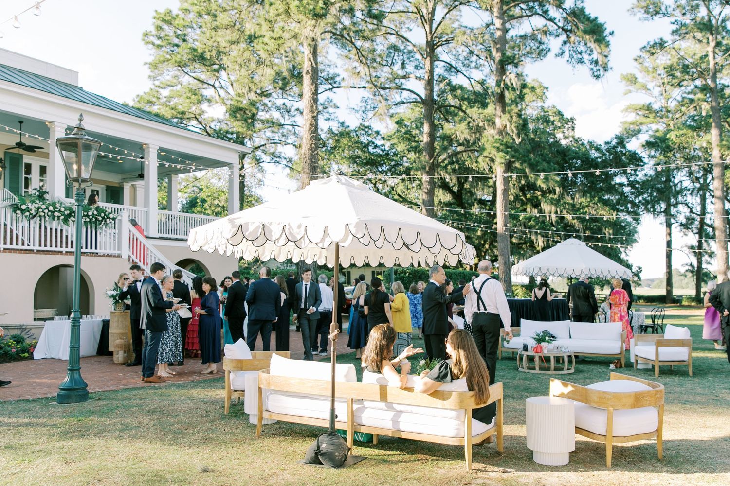 Elegant outdoor wedding reception with white market umbrellas and lounge seating setup on a sunny lawn near a reception house.