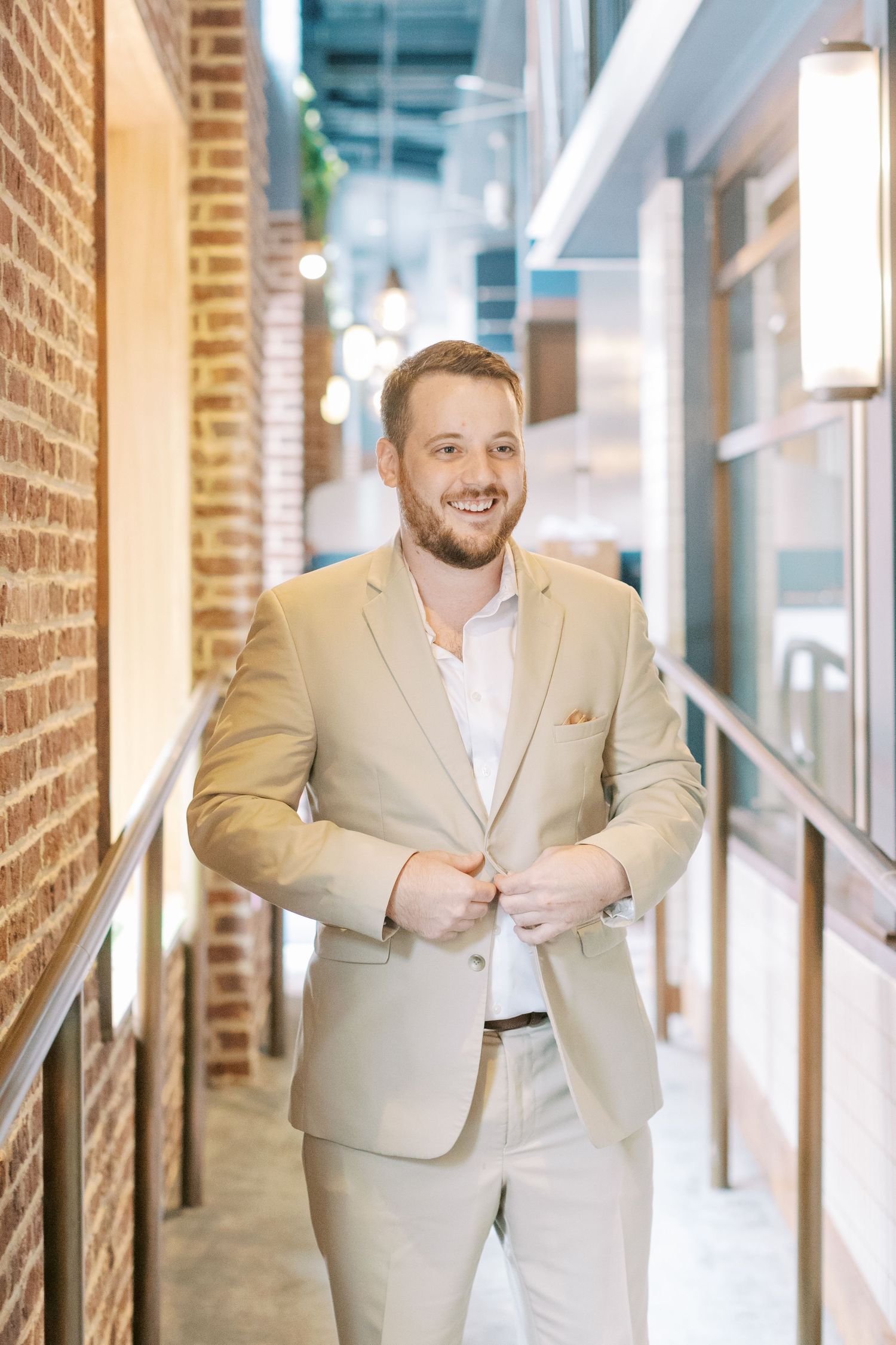 Professional in a tan suit smiles warmly while standing in a modern office hallway with exposed brick walls.