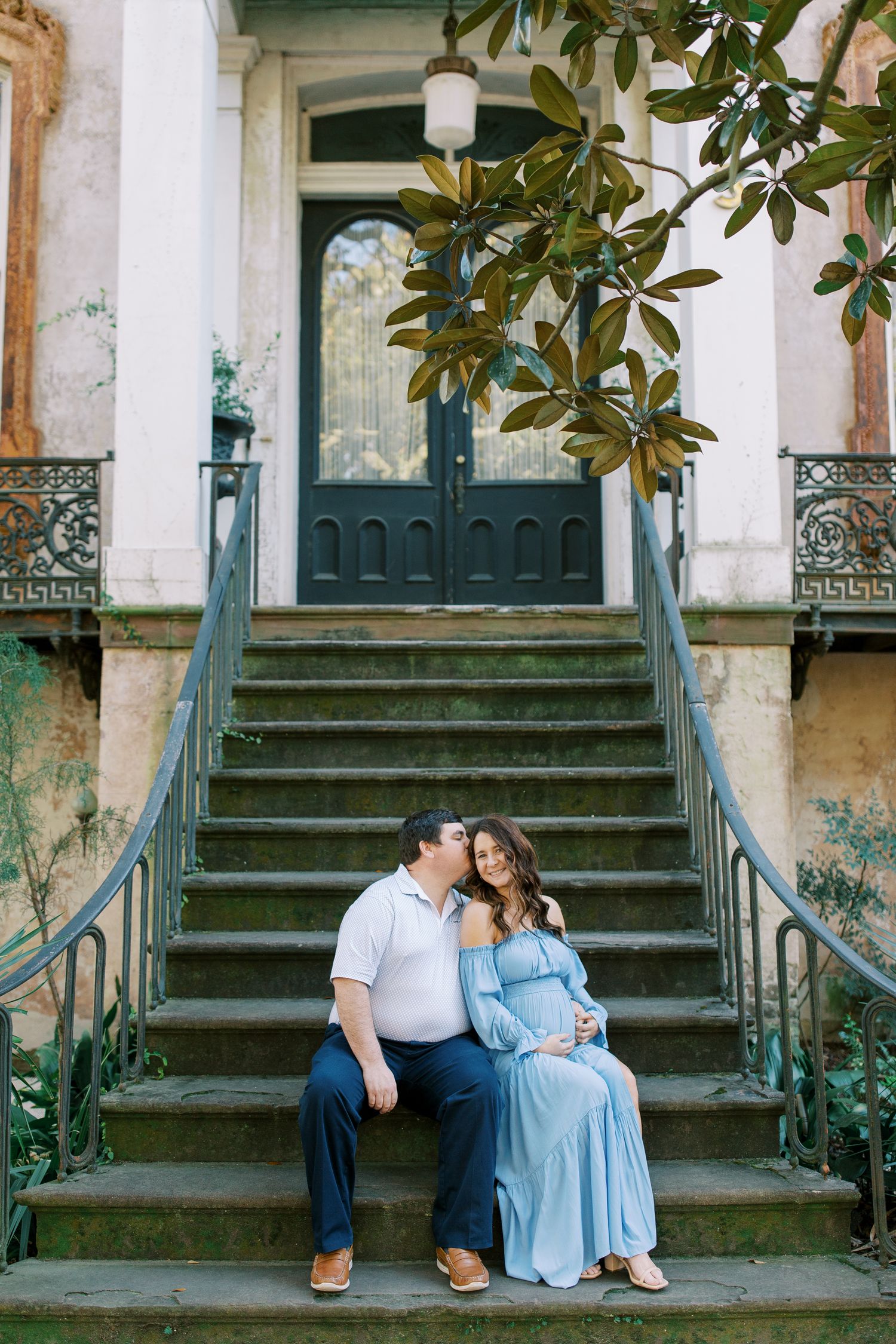 A couple sits together on historic stone steps with ornate iron railings and a grand entrance door.