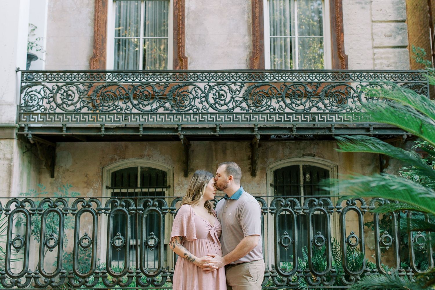 A couple embraces on a wrought iron balcony of a historic building with ornate architectural details and palm fronds.