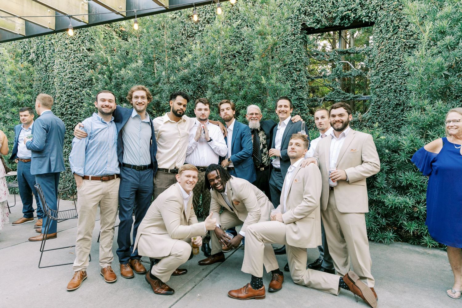 Groomsmen in light suits gather for outdoor photo with dog at garden wedding venue.