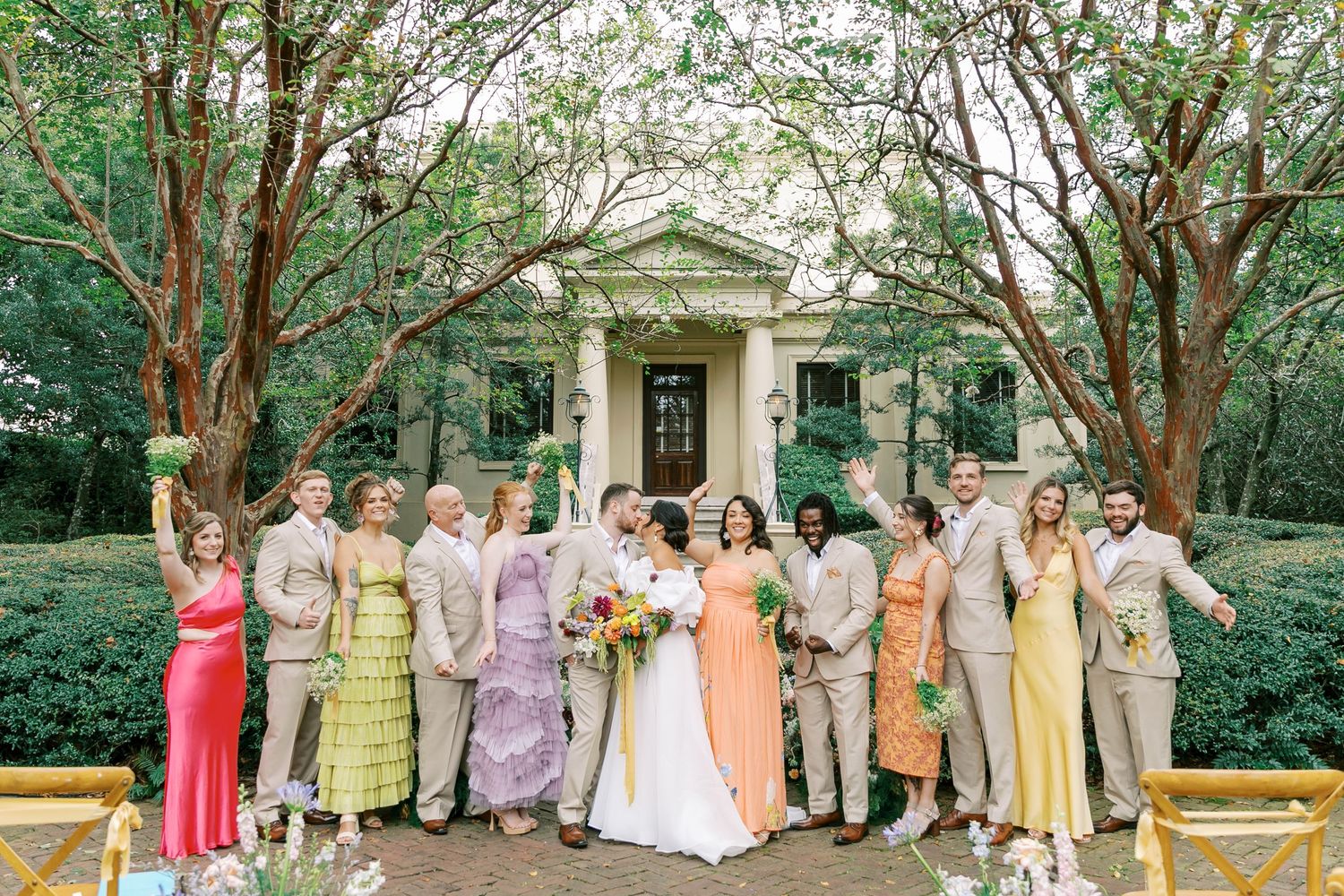 A wedding party wearing pastel dresses poses in front of a grand stone entrance with green trees overhead.