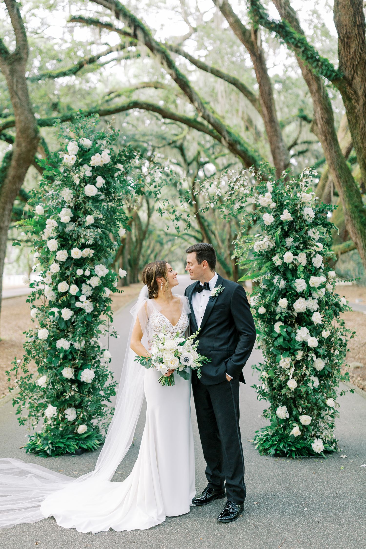 A wedding couple stands beneath a beautiful floral arch made of white roses and greenery in a tree-lined path.