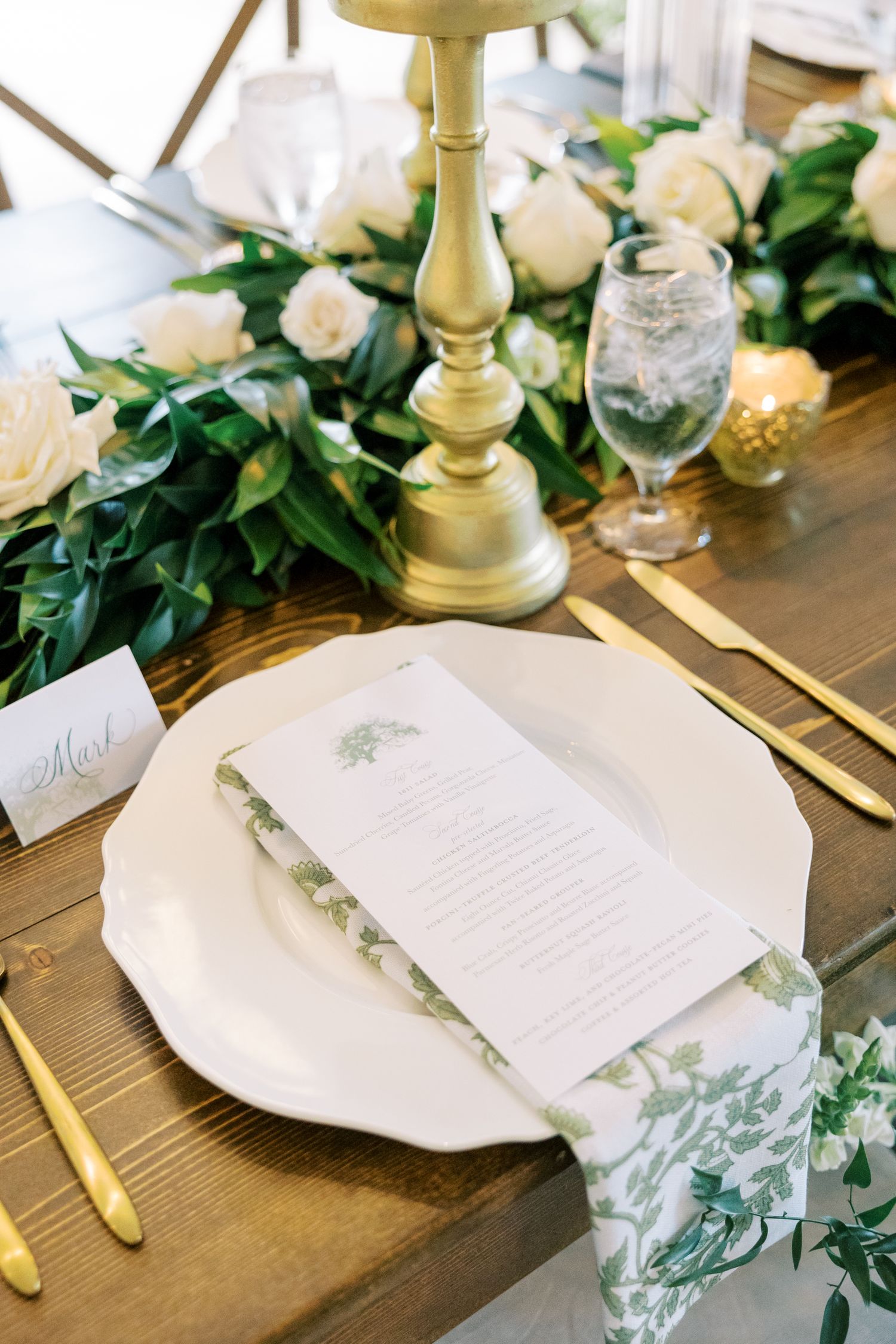 Elegant wedding table setting with white plate, gold candlestick, and white roses on wooden table with greenery.