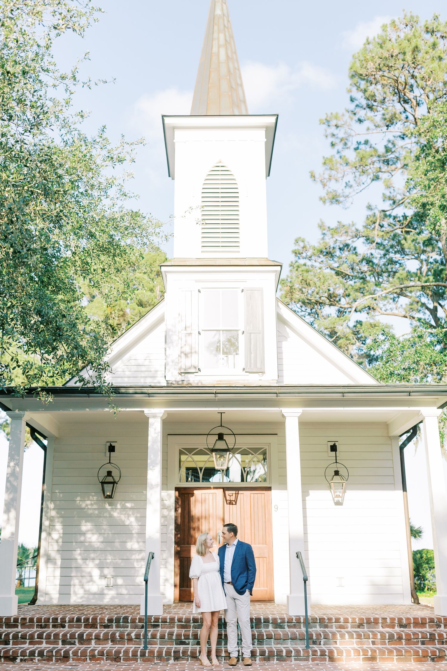 A classic white chapel with tall steeple, brick steps, and hanging lanterns stands surrounded by green trees on a sunny day.
