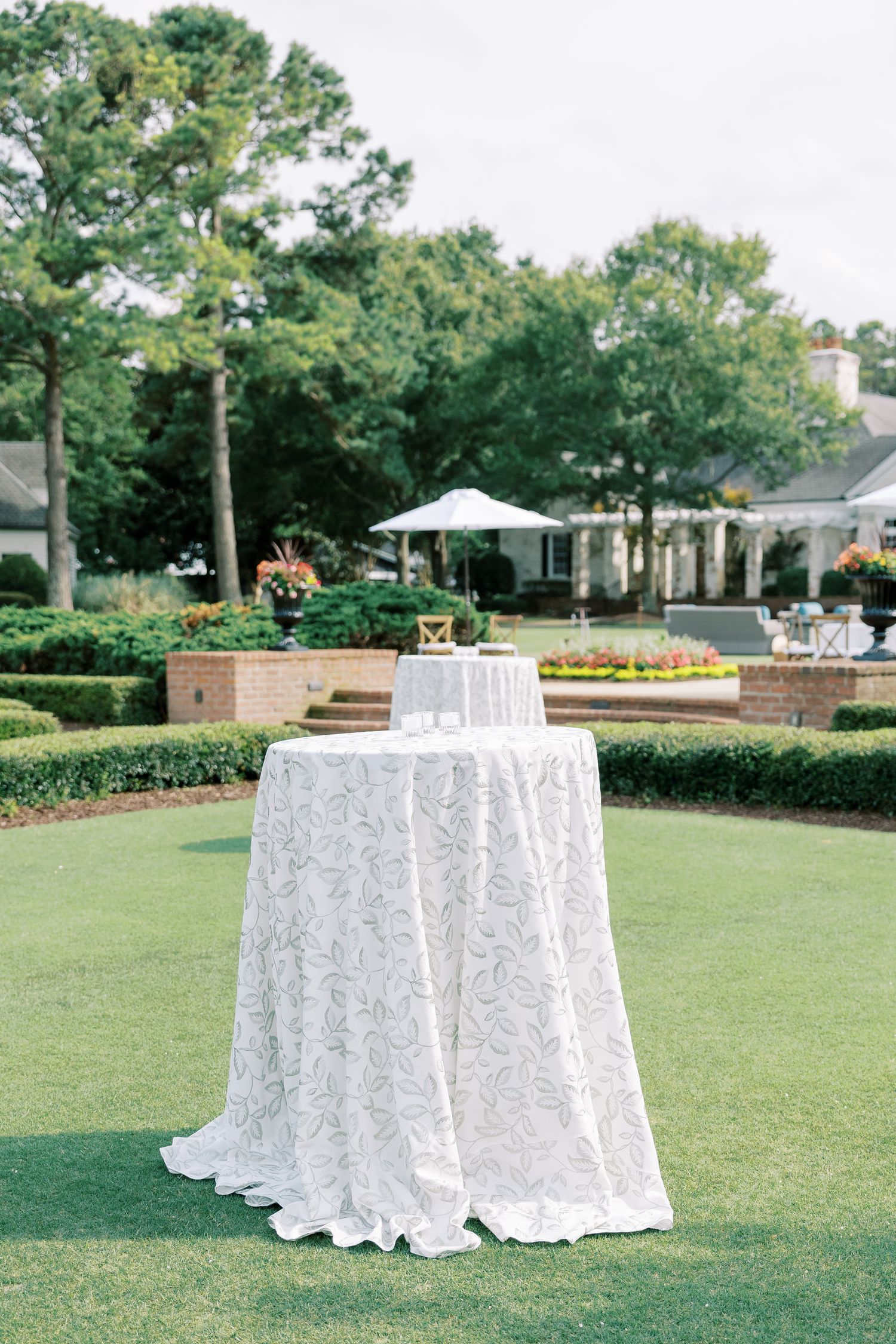 A white lace tablecloth-covered cocktail table sits on manicured lawn with garden hedges and umbrellas in the background.