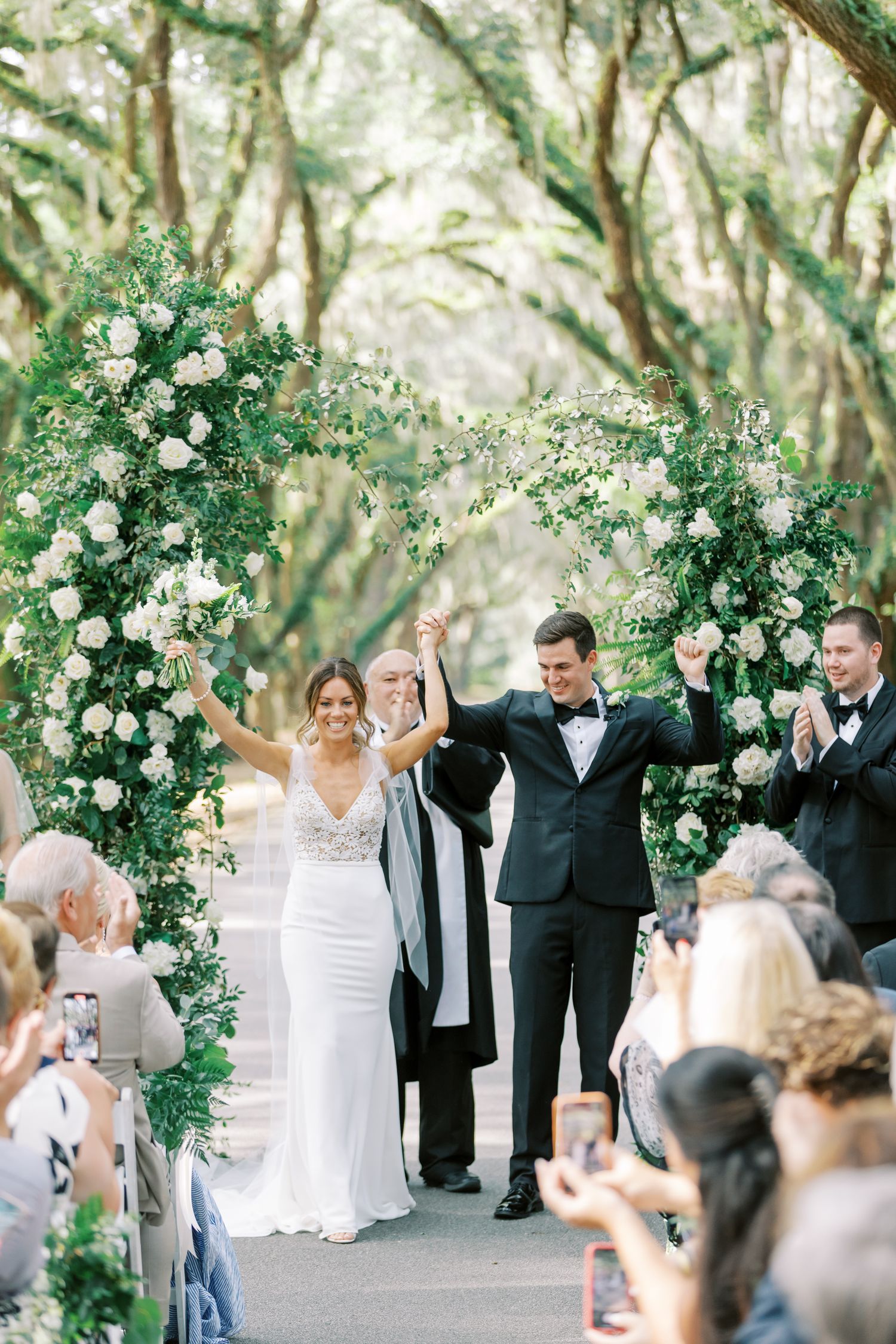 Newlyweds celebrate under an ornate white floral arch while guests capture the joyful moment on their phones.