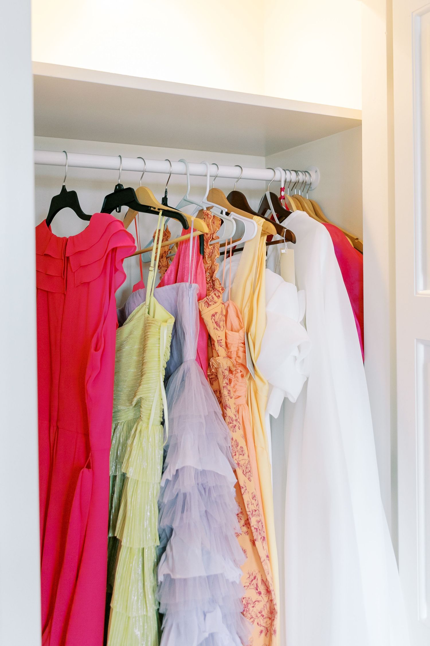 Colorful dresses and formal gowns hanging neatly in a white closet, including pink, green, purple and white garments.
