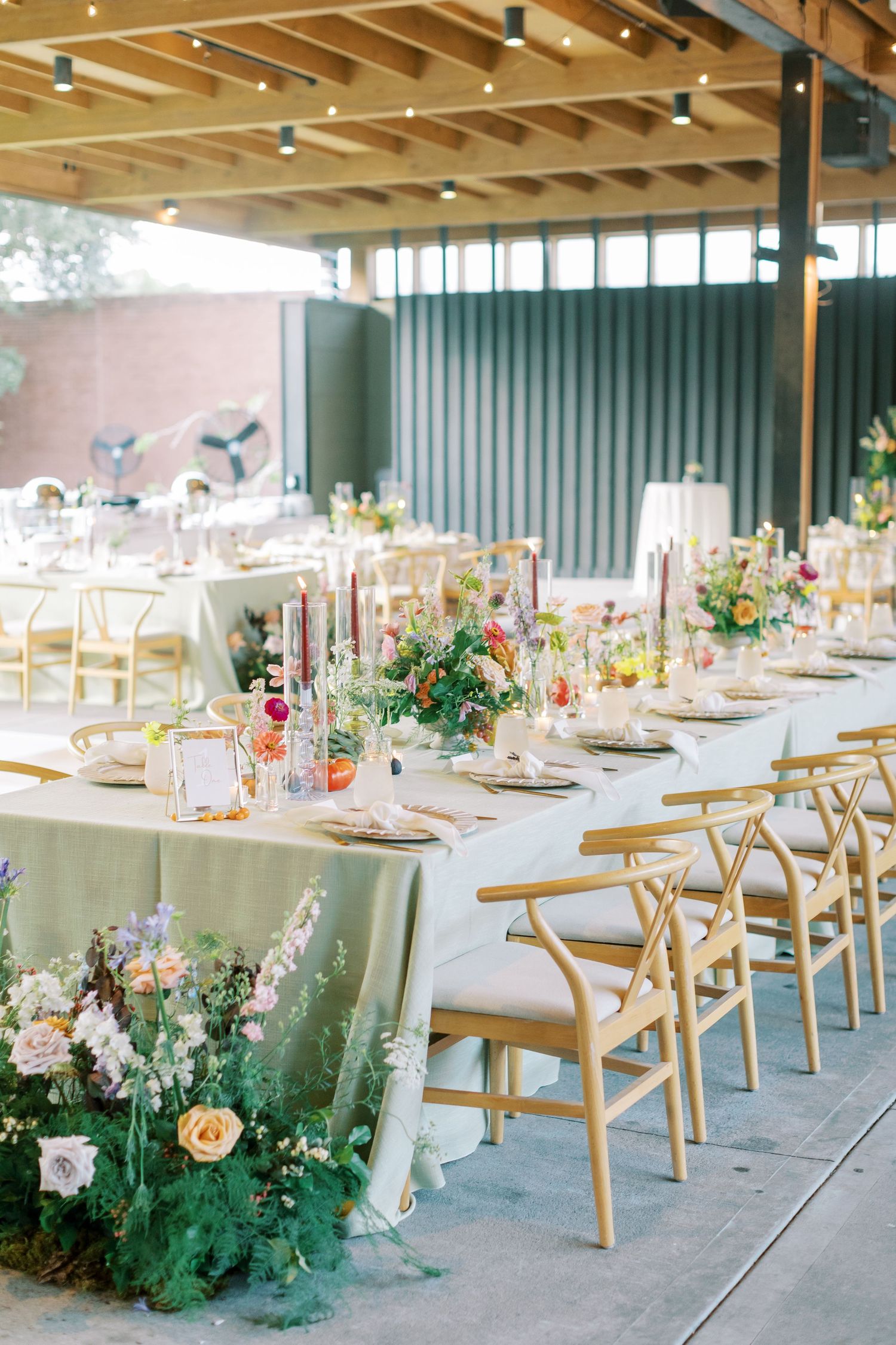 Elegant wedding reception table setting with delicate floral arrangements and gold chairs under wooden beamed ceiling.