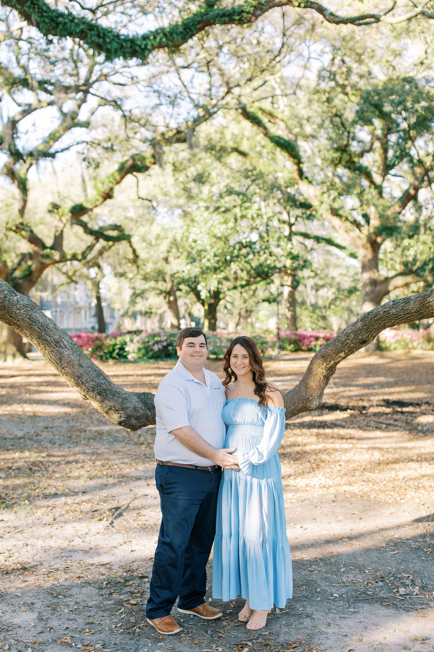 A couple poses under sprawling oak trees in a sunlit park setting.