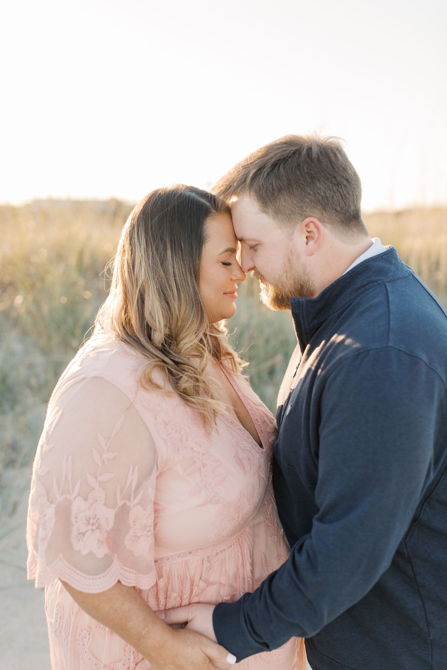 Romantic couple in soft pink dress and navy sweater share an intimate moment in a sunlit field at golden hour.