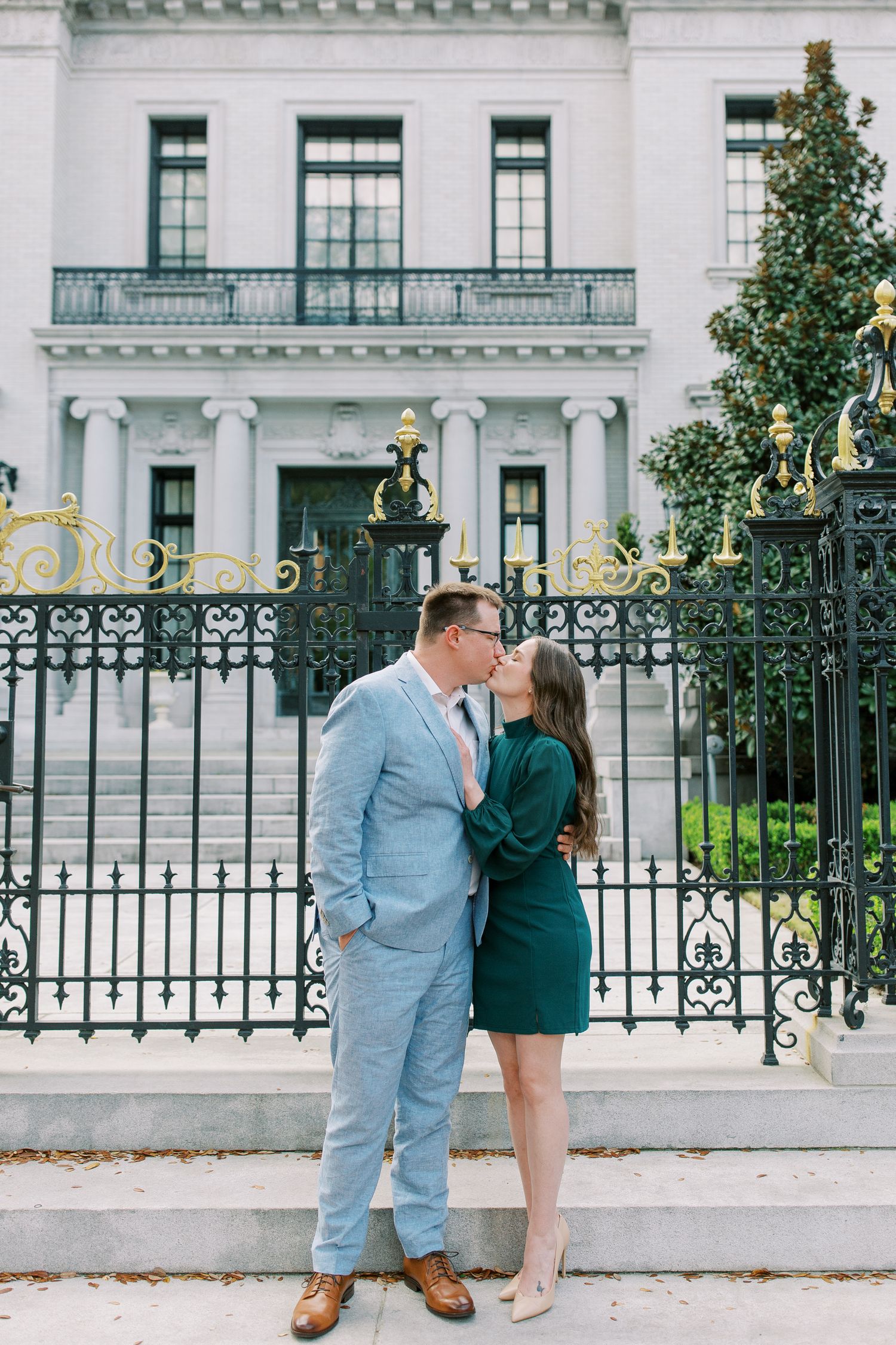 A couple shares a romantic moment in front of an elegant white mansion with ornate iron gates.