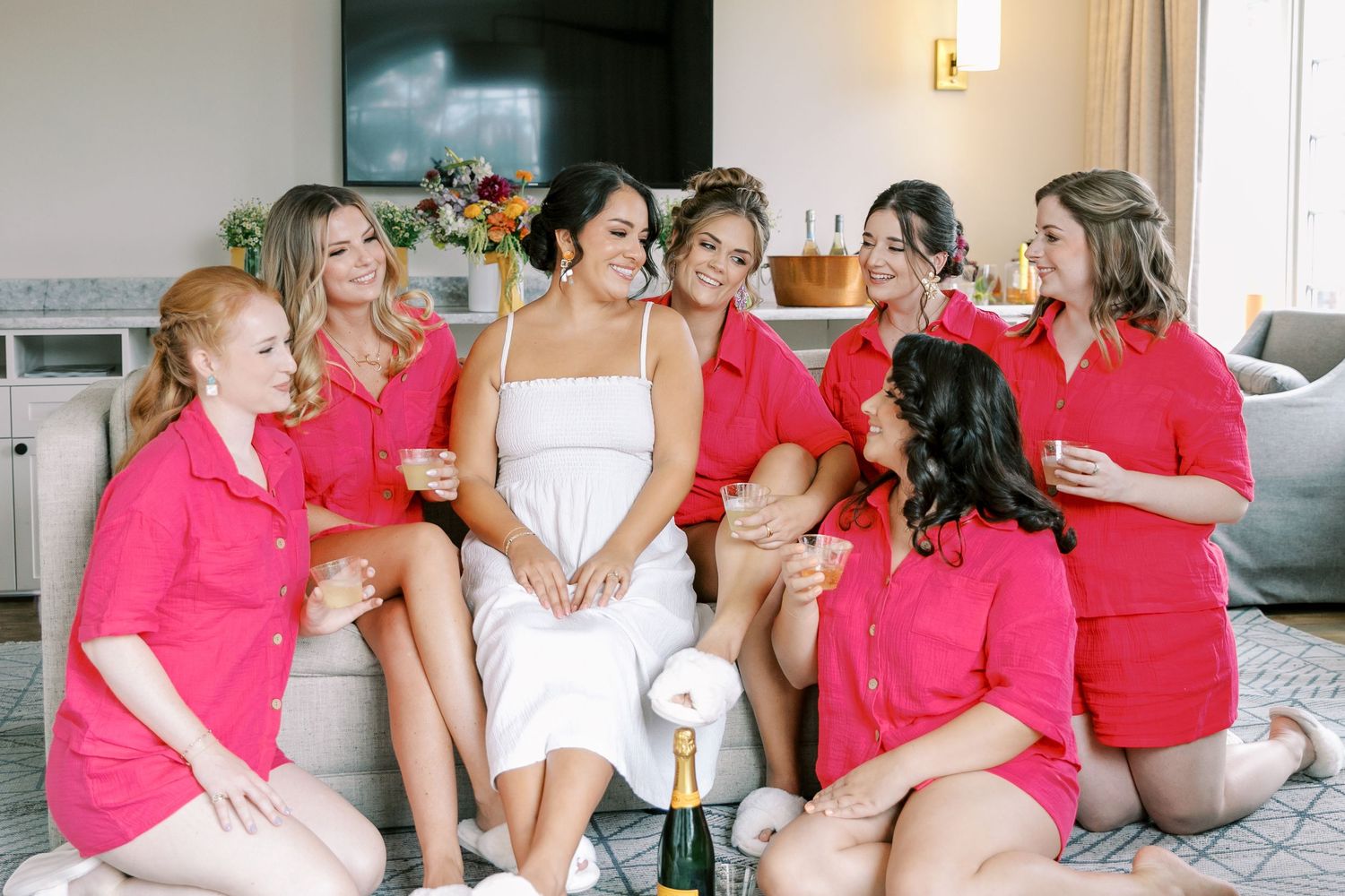 A group wearing pink and white outfits sits together on a couch during what appears to be a wedding preparation event.