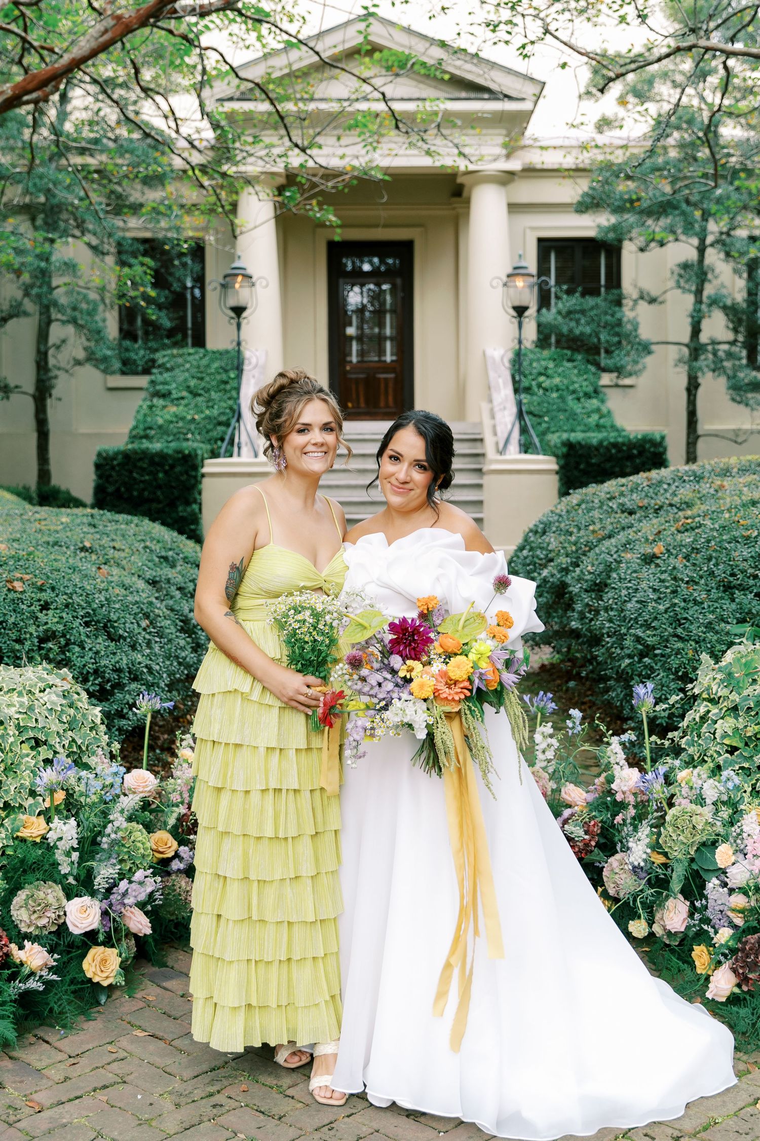 Wedding party members in yellow and white dresses pose together on garden grounds.