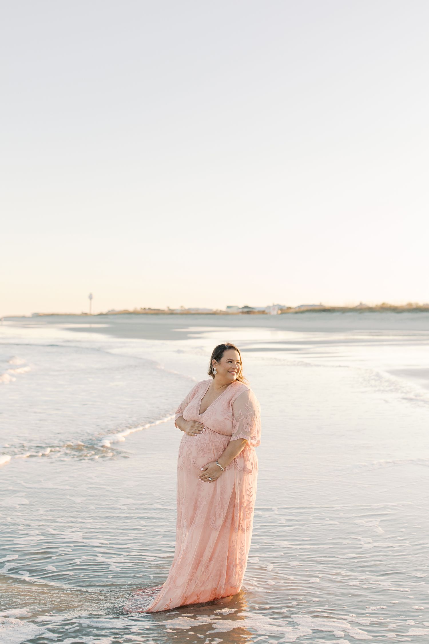 A woman who is pregnant in a flowing pink dress stands at the edge of the ocean during sunset.