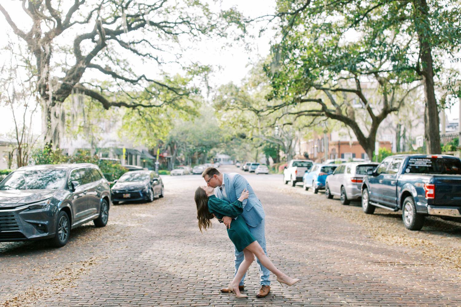 A romantic dip kiss on a tree-lined street with parked cars in the background.